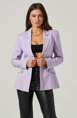 A woman is dressed in a light purple blazer with a notched lapel collar and long sleeves. Underneath, she wears a black crop top, exposing her midriff. She pairs the outfit with high-waisted black leather pants. She accessorizes with large gold hoop earrings, a chunky gold chain necklace, and multiple gold rings.