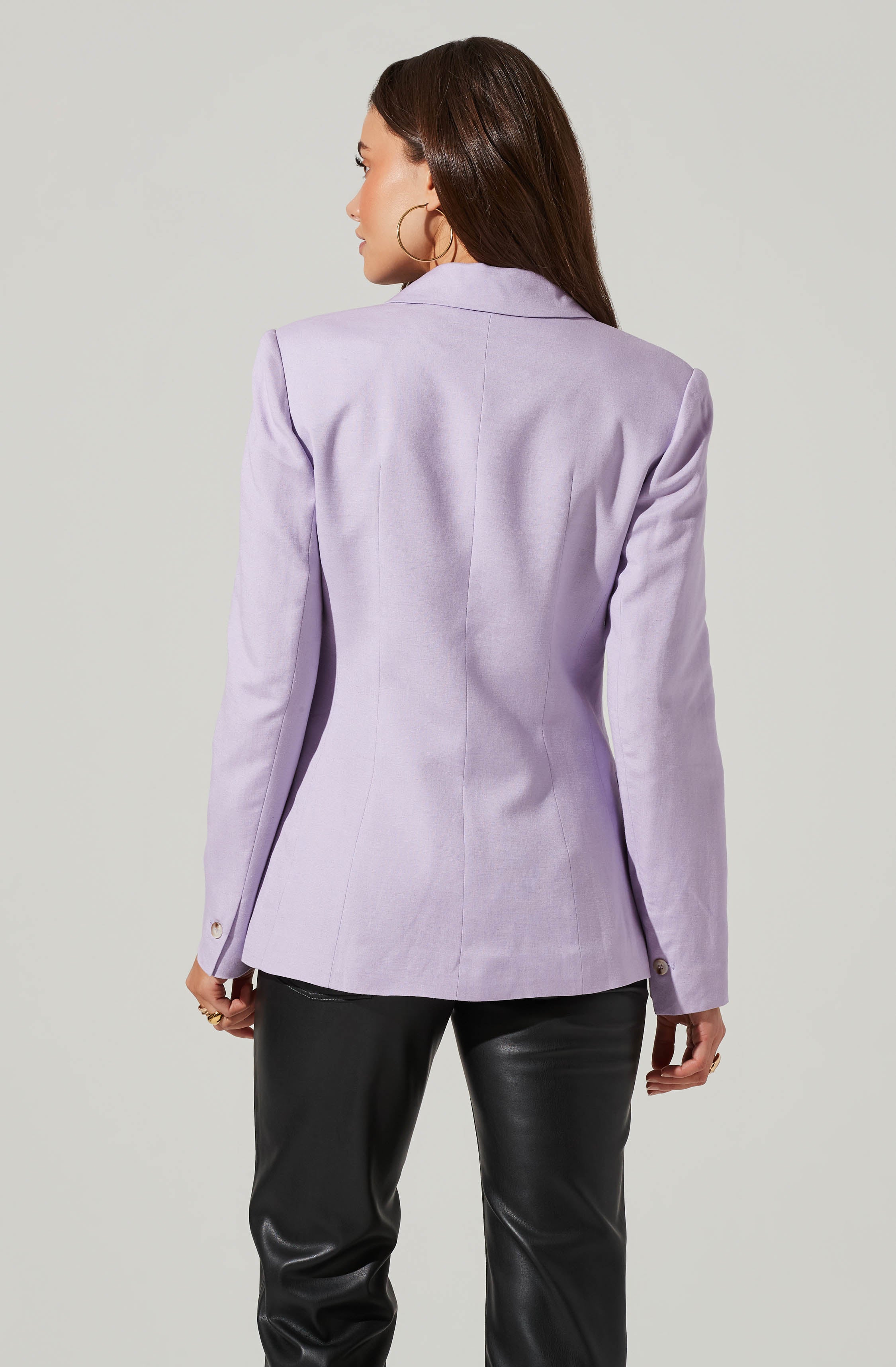 A woman is wearing a light purple blazer with a tailored fit, featuring a notched collar, structured shoulders, and long sleeves with a button at each cuff. She pairs it with black, shiny, leather-like pants. She has long, dark brown hair and is accessorized with large gold hoop earrings and a gold ring on her right hand.