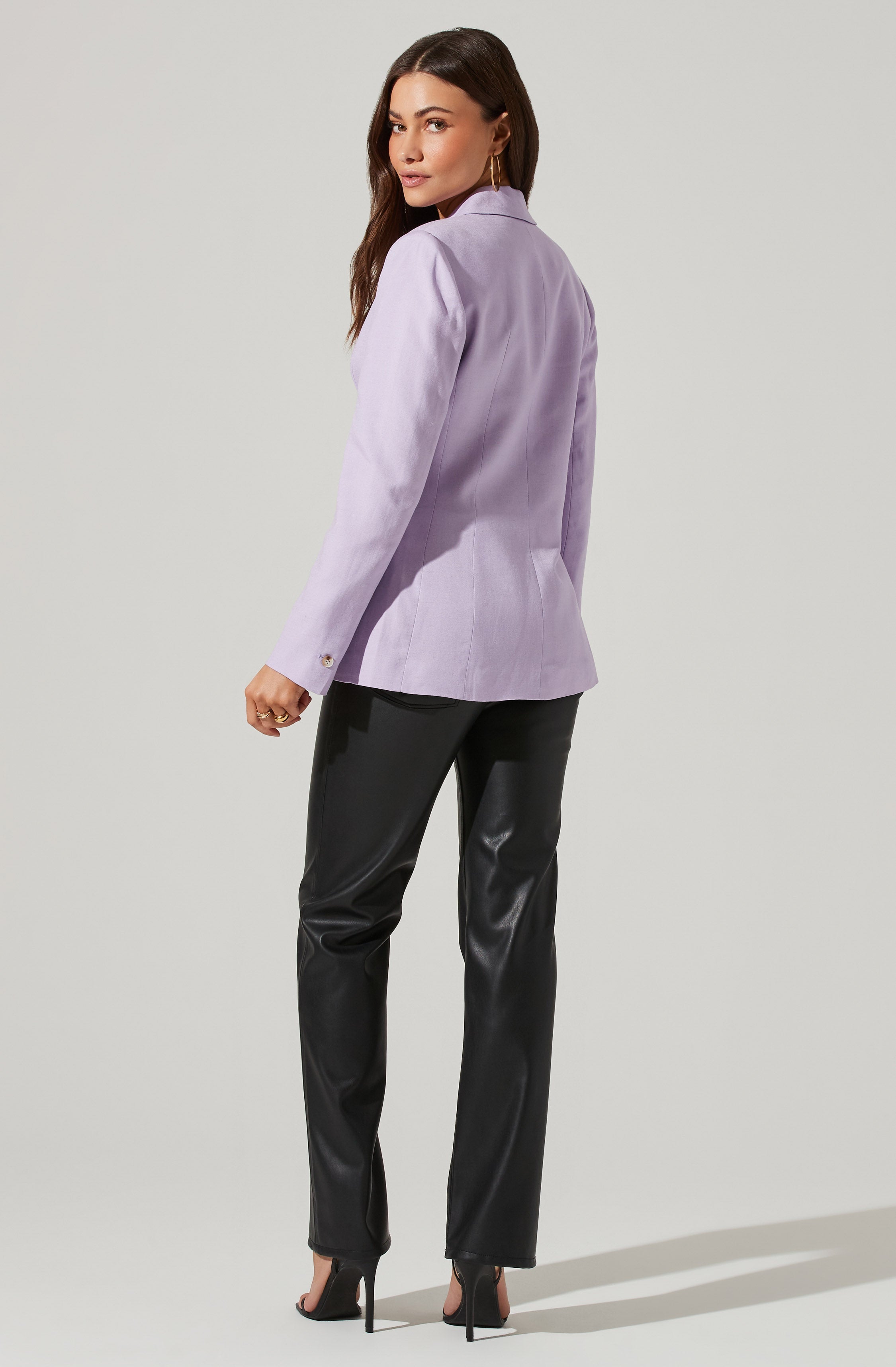 A woman is wearing a light purple blazer with a tailored fit, featuring a notched lapel collar and long sleeves with buttoned cuffs. She pairs it with black, shiny, faux leather pants and black high-heeled open-toe shoes.