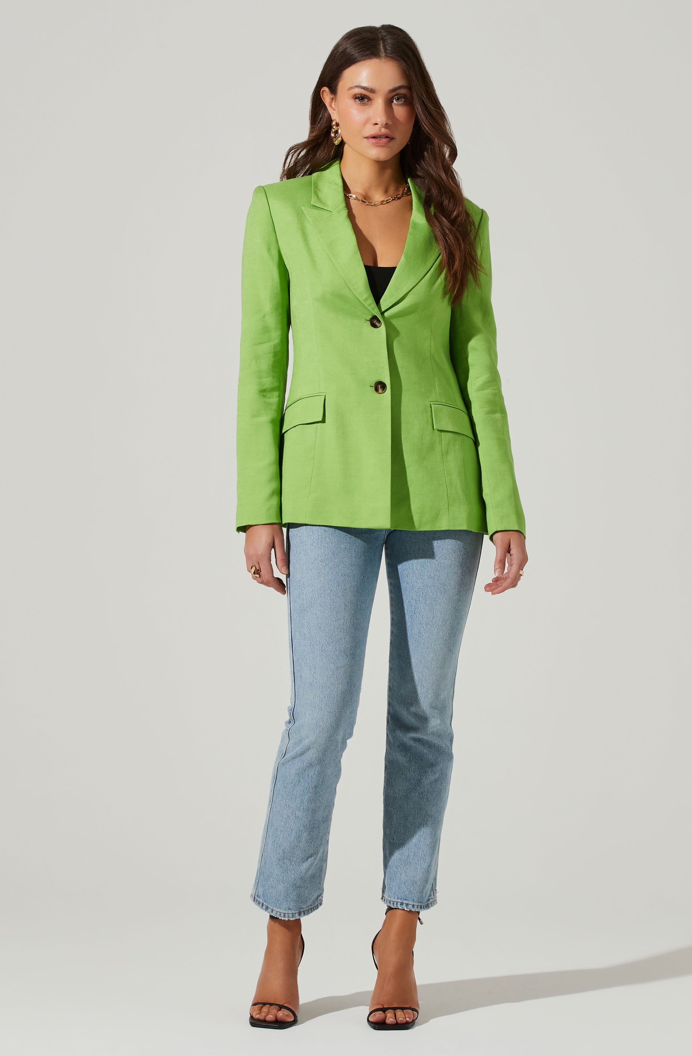 A woman is wearing a light green blazer that features a notched lapel collar, two black buttons down the front, and flap pockets on each side. The blazer is tailored and fitted, giving it a polished appearance. She pairs it with light blue straight-leg jeans and black open-toe high-heeled sandals. The outfit is accessorized with large earrings, a gold chain necklace, and a ring.
