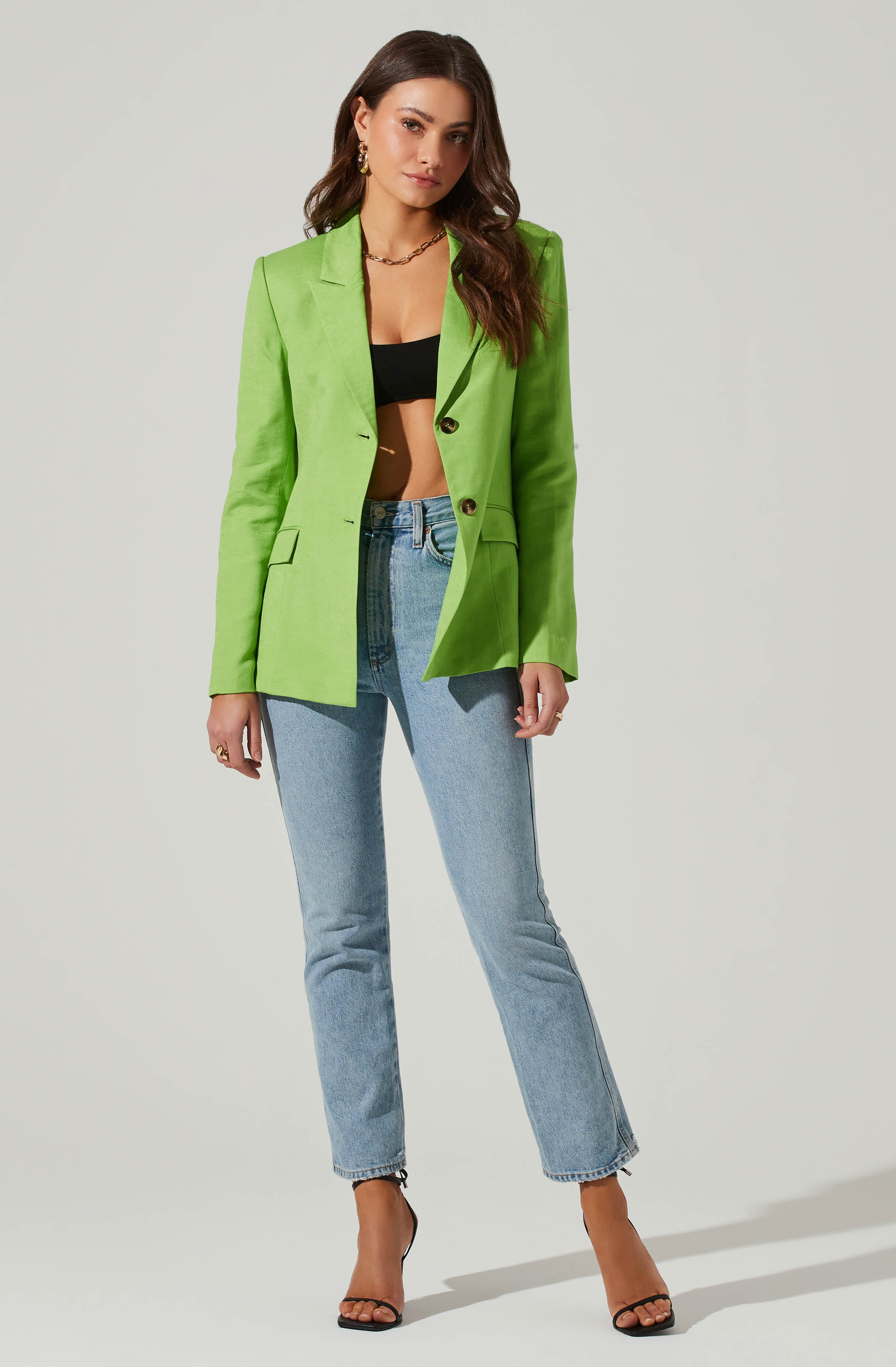 A woman is wearing a bright green blazer with gold buttons, paired with light blue high-waisted jeans. Underneath the blazer, she has on a black crop top. She is accessorized with gold hoop earrings, a gold chain necklace, and a ring. She is also wearing black high-heeled sandals.