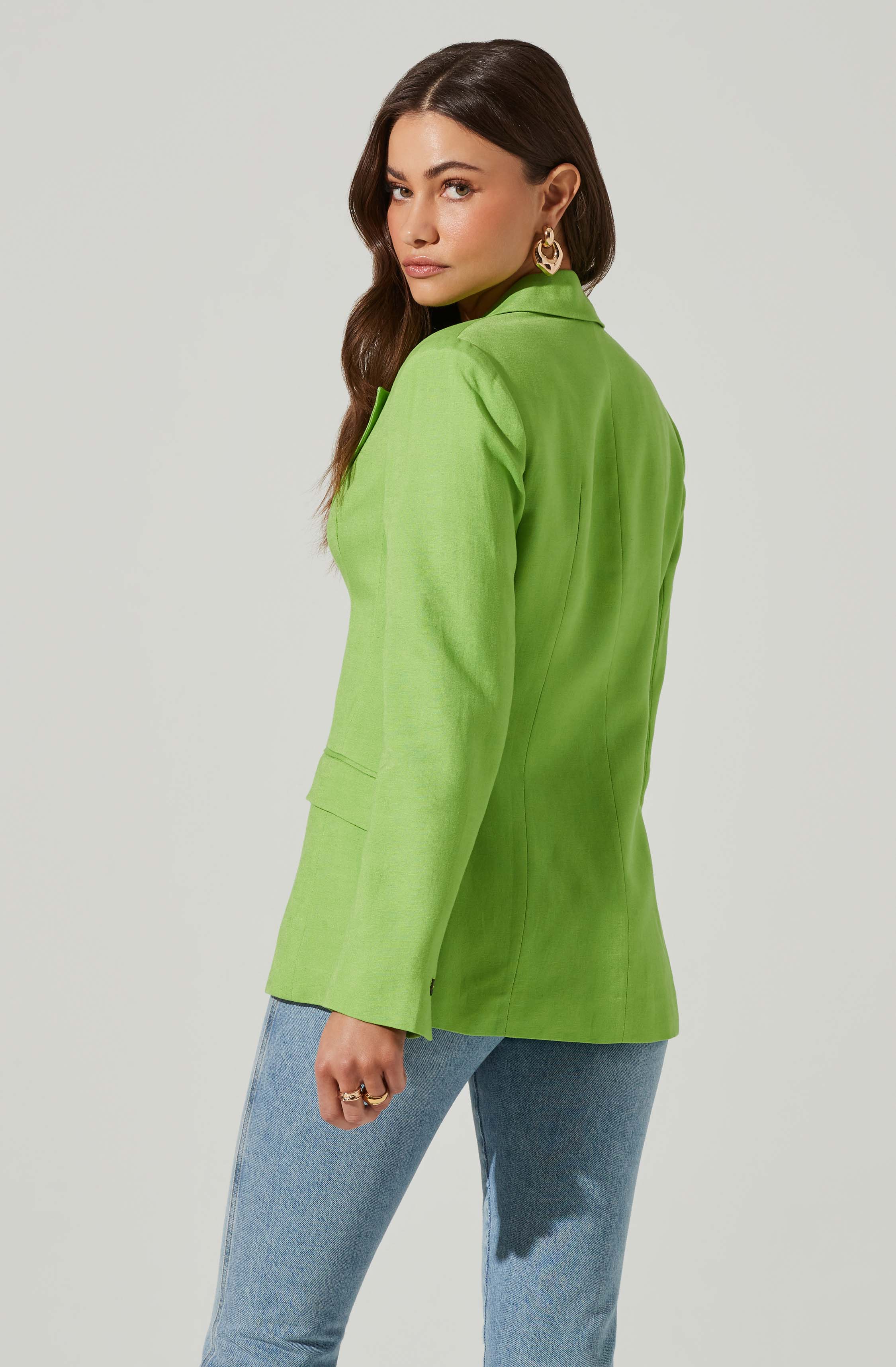 A woman is wearing a bright green blazer with a tailored fit, featuring a notched lapel and long sleeves. She pairs it with light blue denim jeans. She has long, dark brown hair and is accessorized with gold hoop earrings and multiple rings on her right hand.