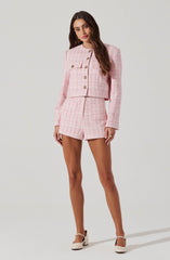 A woman is wearing a light pink, textured, matching two-piece outfit. The top is a cropped, long-sleeve jacket with a round collar and gold buttons down the front, featuring a flap pocket on the left side. She pairs it with high-waisted matching shorts. She is also wearing white, ankle-strap high heels.