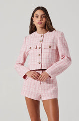 A woman is wearing a light pink, textured, tweed-like suit consisting of a cropped blazer and matching high-waisted shorts. The blazer features a round neckline, long sleeves, and decorative gold buttons down the front. It also has two small flap pockets on the chest with gold buttons.