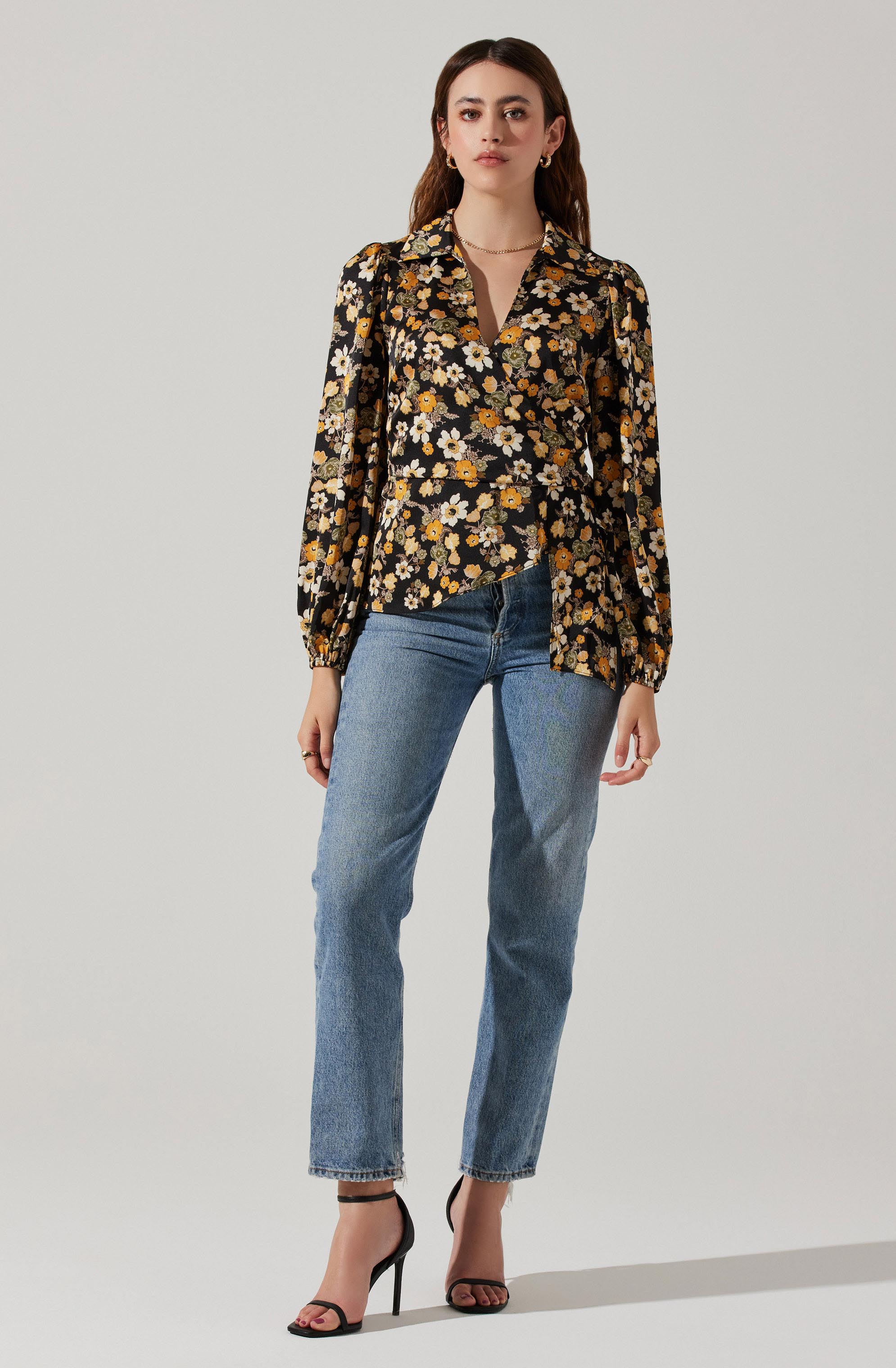 A woman is wearing a long-sleeved, collared blouse with a black background and a floral pattern featuring white, yellow, and orange flowers with green leaves. She has paired the blouse with blue jeans and black high-heeled sandals.