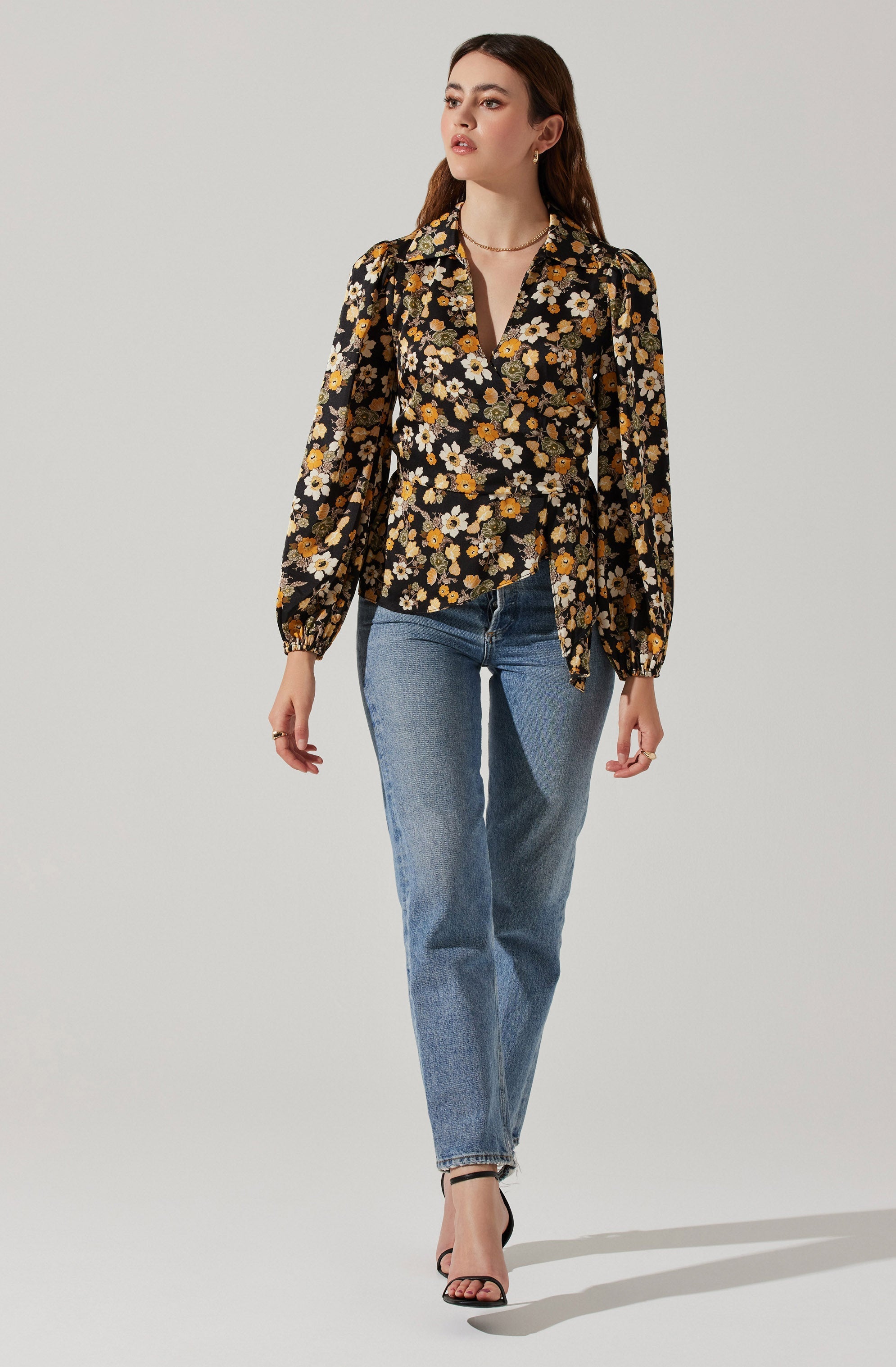 A woman is wearing a long-sleeved, collared blouse with a black background and a yellow, white, and orange floral pattern. She has paired it with high-waisted, light blue jeans and black, open-toe high-heeled sandals. She also has small hoop earrings, a necklace, and rings on her fingers.