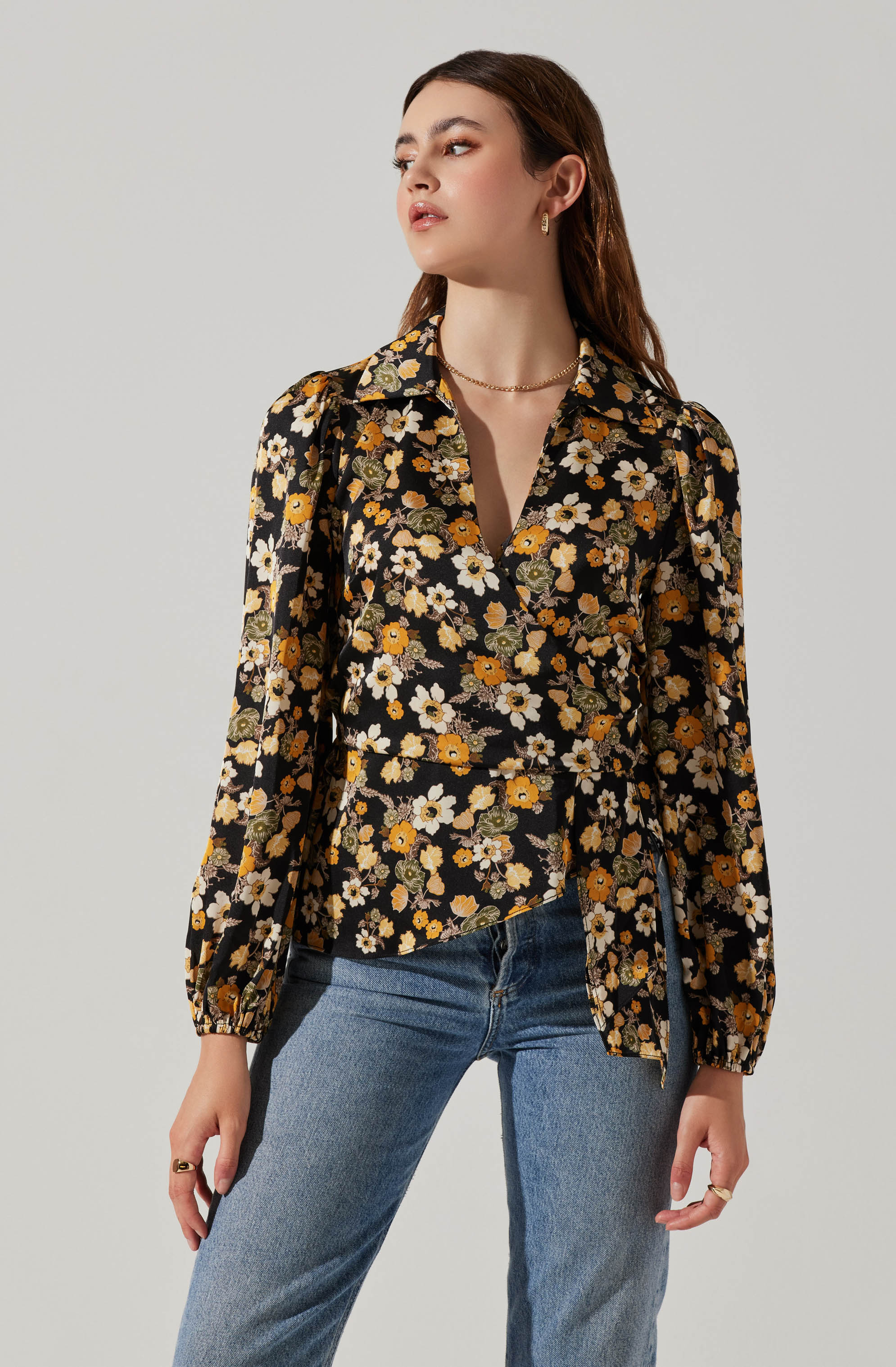 A woman is wearing a long-sleeved, black blouse with a floral pattern consisting of white, orange, and yellow flowers and green leaves. The blouse has a collar and a deep V-neck, with puffed shoulders and gathered cuffs. She pairs it with light blue jeans. She has accessorized with small hoop earrings, a delicate necklace, and rings on her fingers.