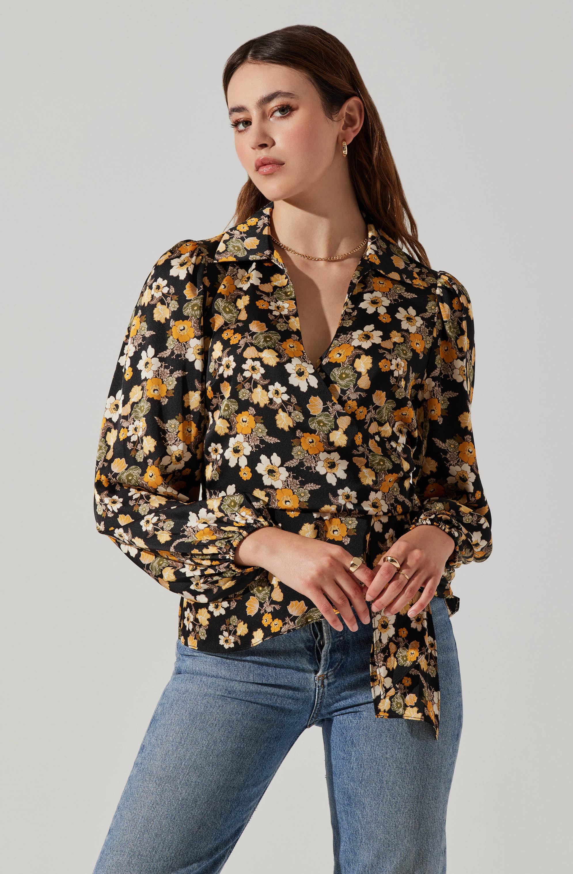 A woman is wearing a black blouse with a vibrant floral pattern in yellow, white, and green, featuring a collar and long, slightly puffed sleeves. She pairs it with medium-wash blue jeans. She accessorizes with small hoop earrings, a gold necklace, and several rings on her fingers.