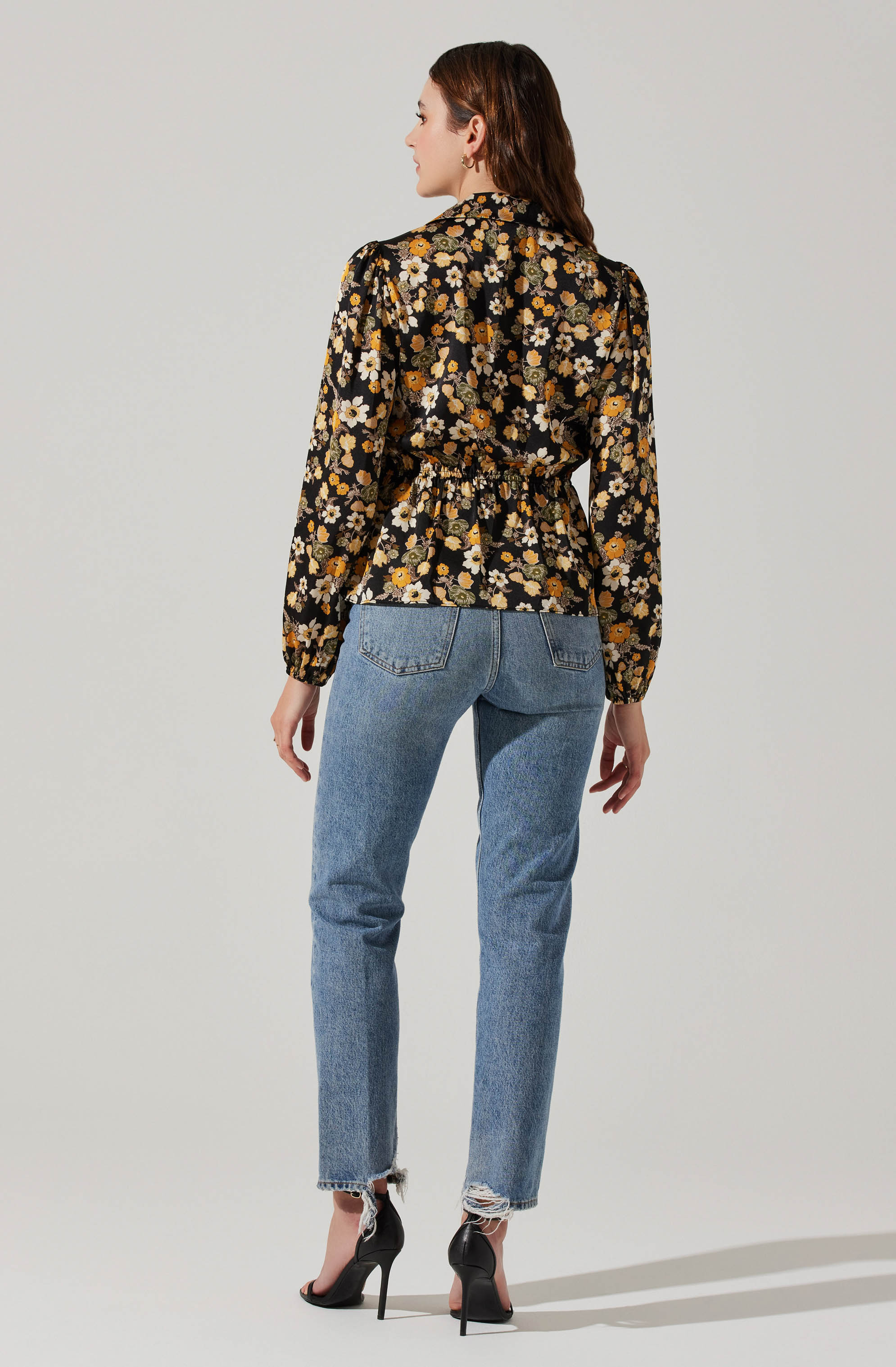 A woman is wearing a black blouse with a floral pattern in shades of yellow, white, and beige. The blouse has long sleeves with gathered cuffs and a cinched waist. She pairs it with light wash, high-waisted jeans that have a frayed hem at the ankles. She is also wearing black high-heeled shoes.