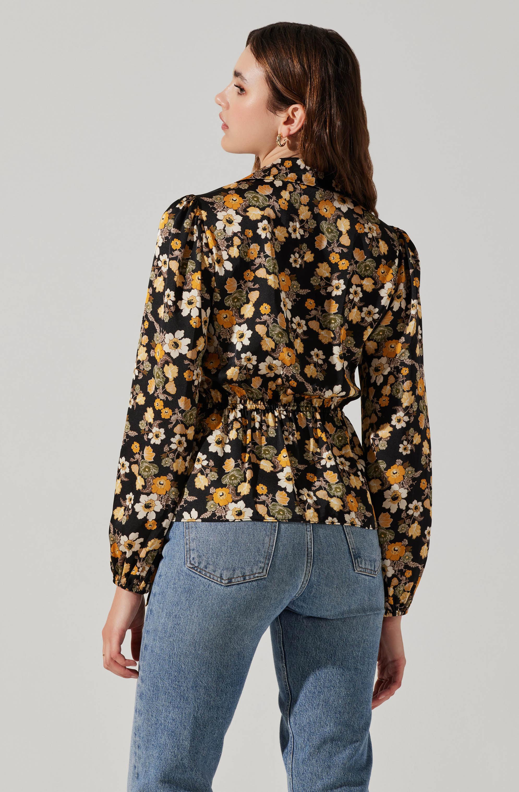 A woman is wearing a black floral blouse with long sleeves, featuring a pattern of yellow, white, and orange flowers with green leaves. The blouse has a gathered, elastic waistband and padded shoulders. She is also wearing light blue denim jeans. The woman has shoulder-length dark brown hair and is wearing small gold hoop earrings.