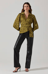 A woman is wearing a silky, olive-green blouse with a black abstract floral pattern, featuring a collar, a deep V-neckline, and puffed long sleeves that are gathered at the cuffs. The blouse is cinched at the waist with a matching fabric belt tied in a knot. She pairs it with high-waisted, black, faux leather wide-leg pants and black high-heeled sandals with thin straps. She has gold hoop earrings and her hair is styled in loose waves.