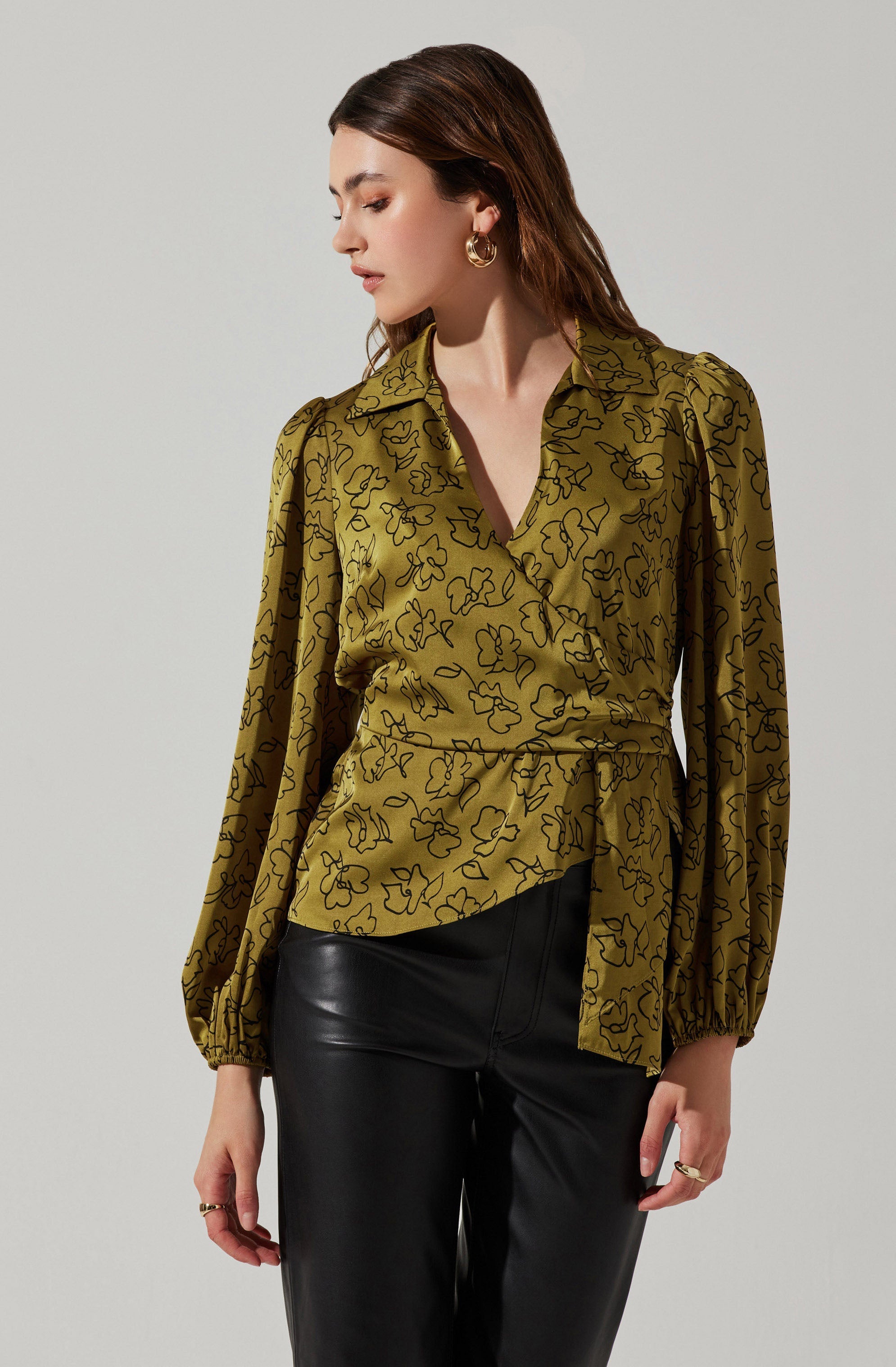 A woman is wearing a mustard yellow blouse with a black abstract floral pattern, featuring a collar, long puffed sleeves with elastic cuffs, and a tied waist. She is paired with black leather pants. She has medium-length brown hair, gold hoop earrings, and a gold ring on her right hand.