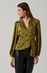 A woman is wearing a mustard yellow blouse with a black abstract floral pattern, featuring a collar, long puffed sleeves with elastic cuffs, and a tied waist. She is paired with black leather pants. She has medium-length brown hair, gold hoop earrings, and a gold ring on her right hand.