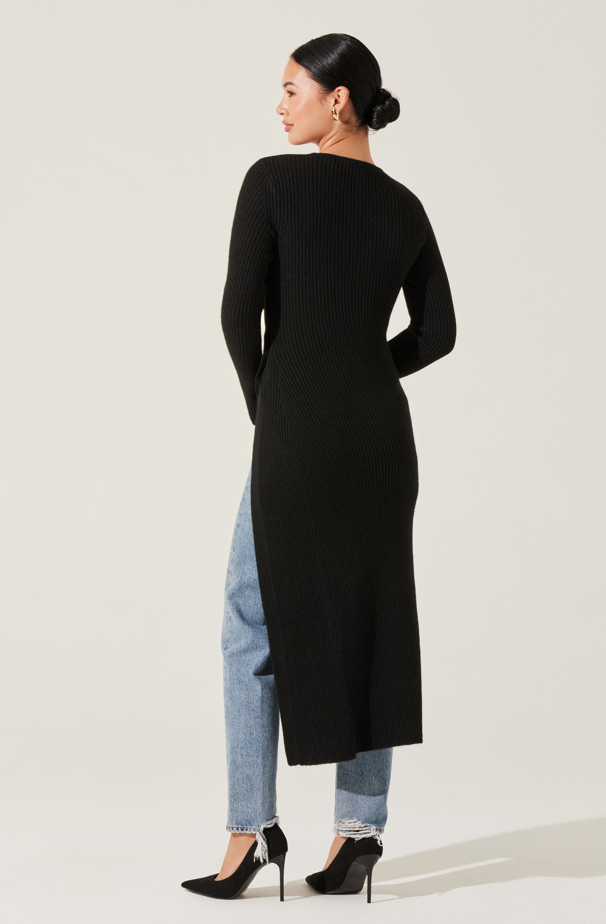 A woman is wearing a long, black, ribbed knit dress with long sleeves that reaches down to her ankles. She has paired the dress with light blue, slightly distressed jeans rolled at the cuffs and black high-heeled shoes with pointed toes and slender heels.