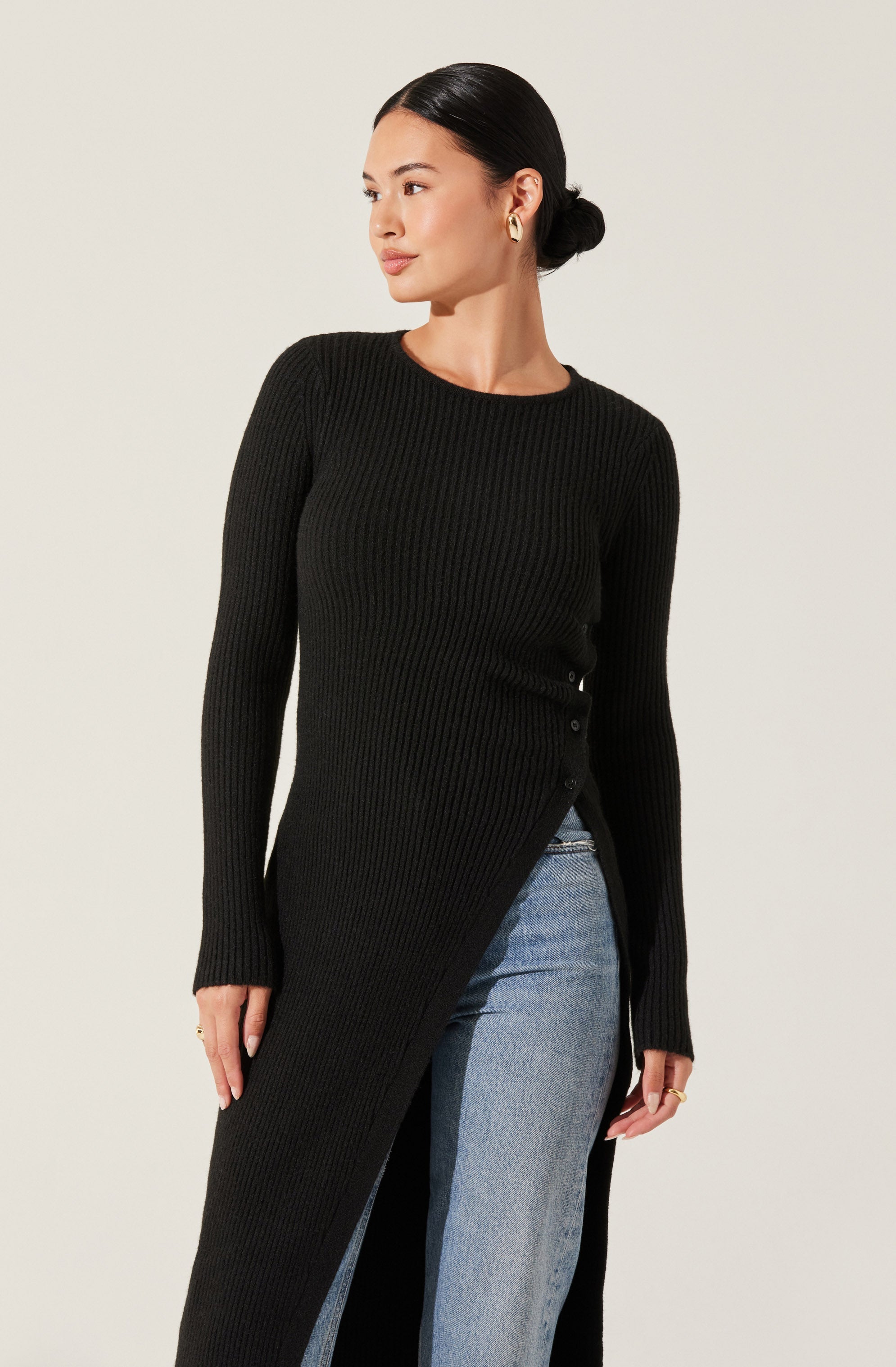 A woman is wearing a long, black, ribbed knit sweater with buttons on the side. She has her hair tied back in a low bun and is accessorizing with gold hoop earrings and rings. She is also wearing light-wash blue jeans.