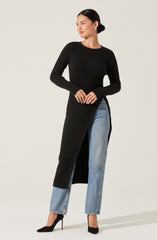 A woman is wearing a long black ribbed knit sweater that extends to her ankles with a side slit. She pairs it with light blue, high-waisted, straight-leg jeans with a slightly faded wash. She completes the outfit with black high-heeled pumps and simple gold hoop earrings.