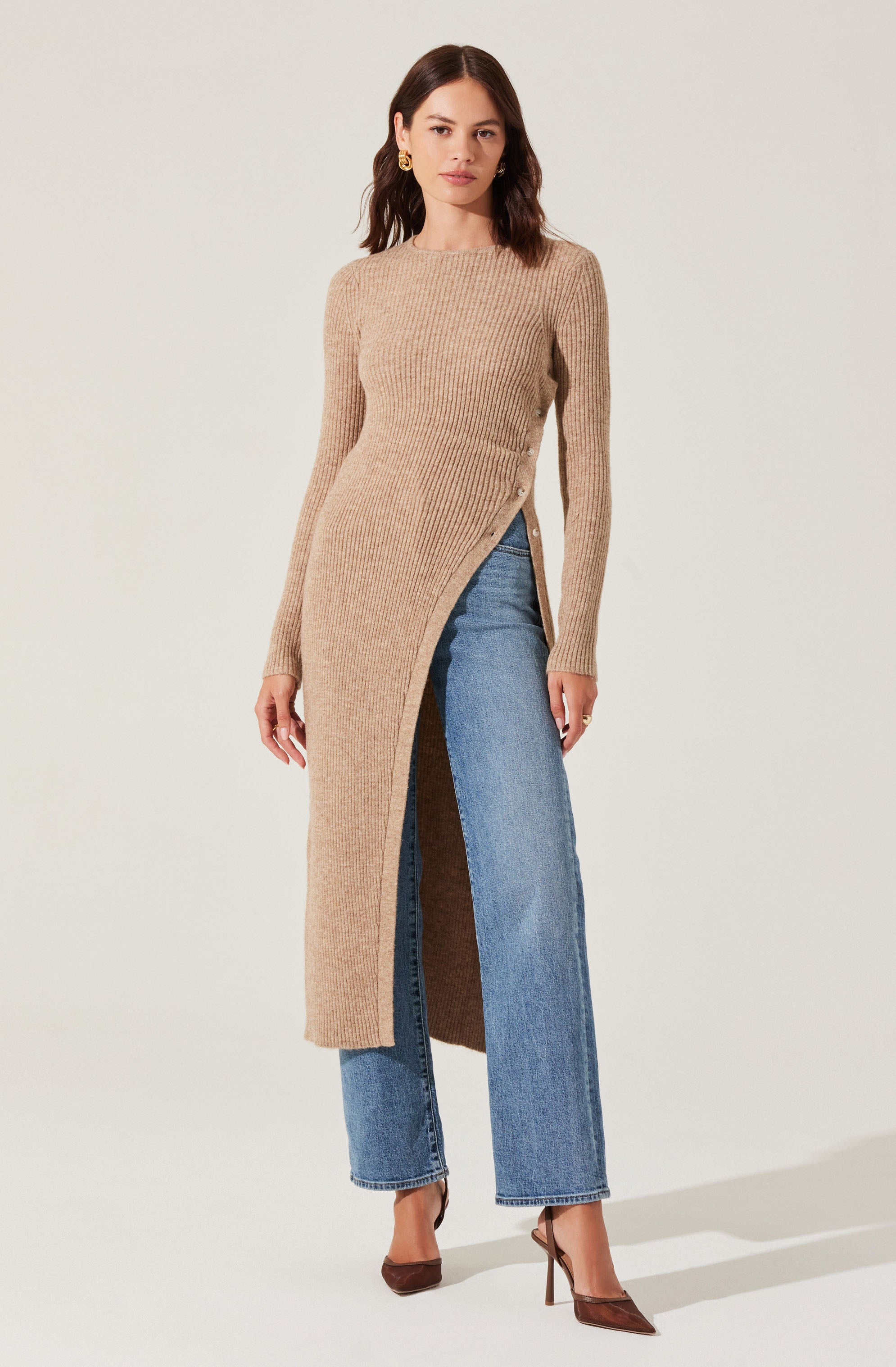 A woman is wearing a beige, ribbed knit sweater dress with a high neckline that features a front slit running from the hem to above the knee. She pairs it with high-waisted, straight-leg blue jeans and brown pointed-toe high heels. She has gold earrings and dark, wavy hair.