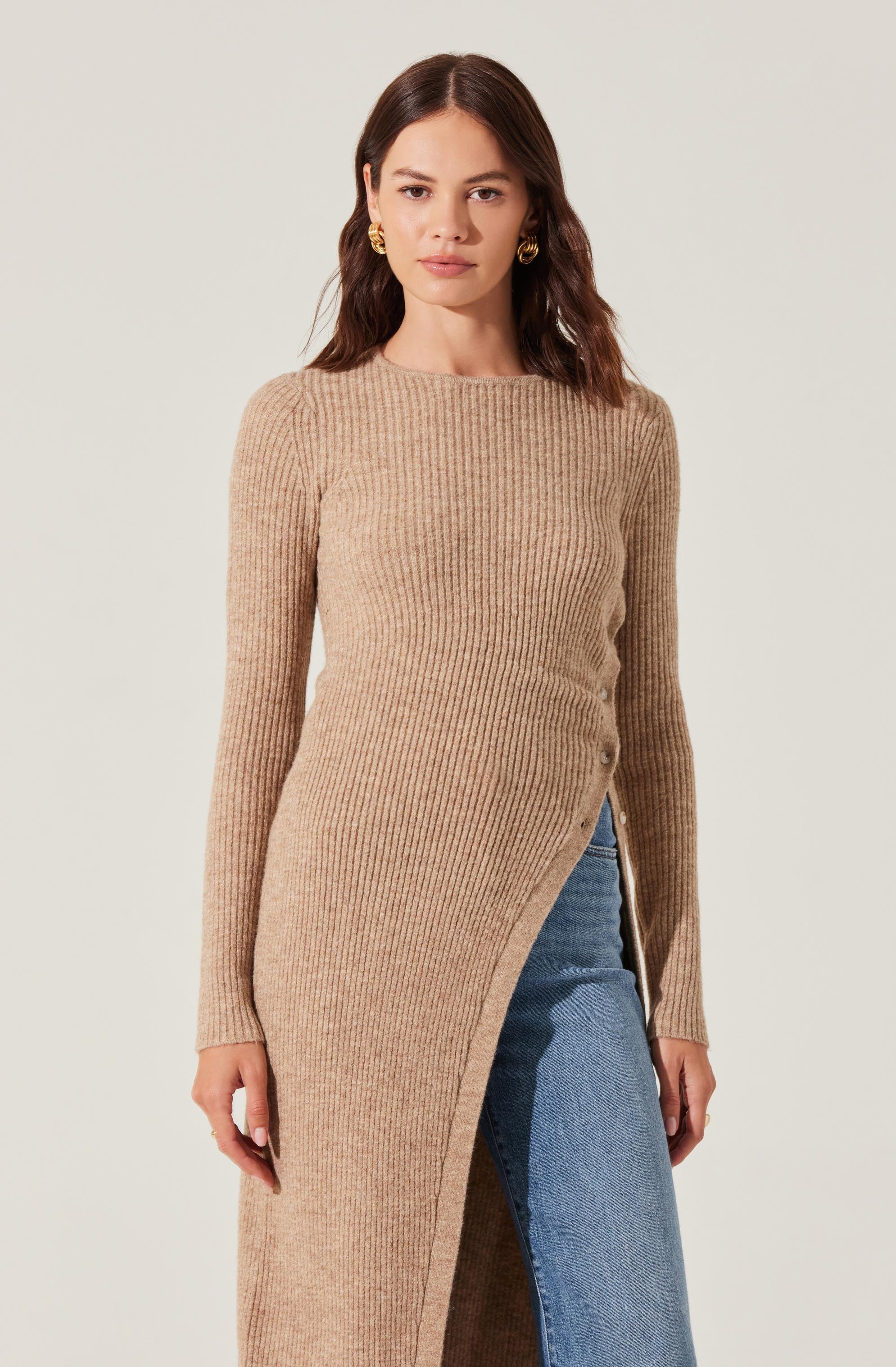 A woman is wearing a long, beige, ribbed knit sweater with an asymmetrical front slit that reveals part of her blue denim skirt. She has long, dark brown hair and is accessorized with large, gold hoop earrings.