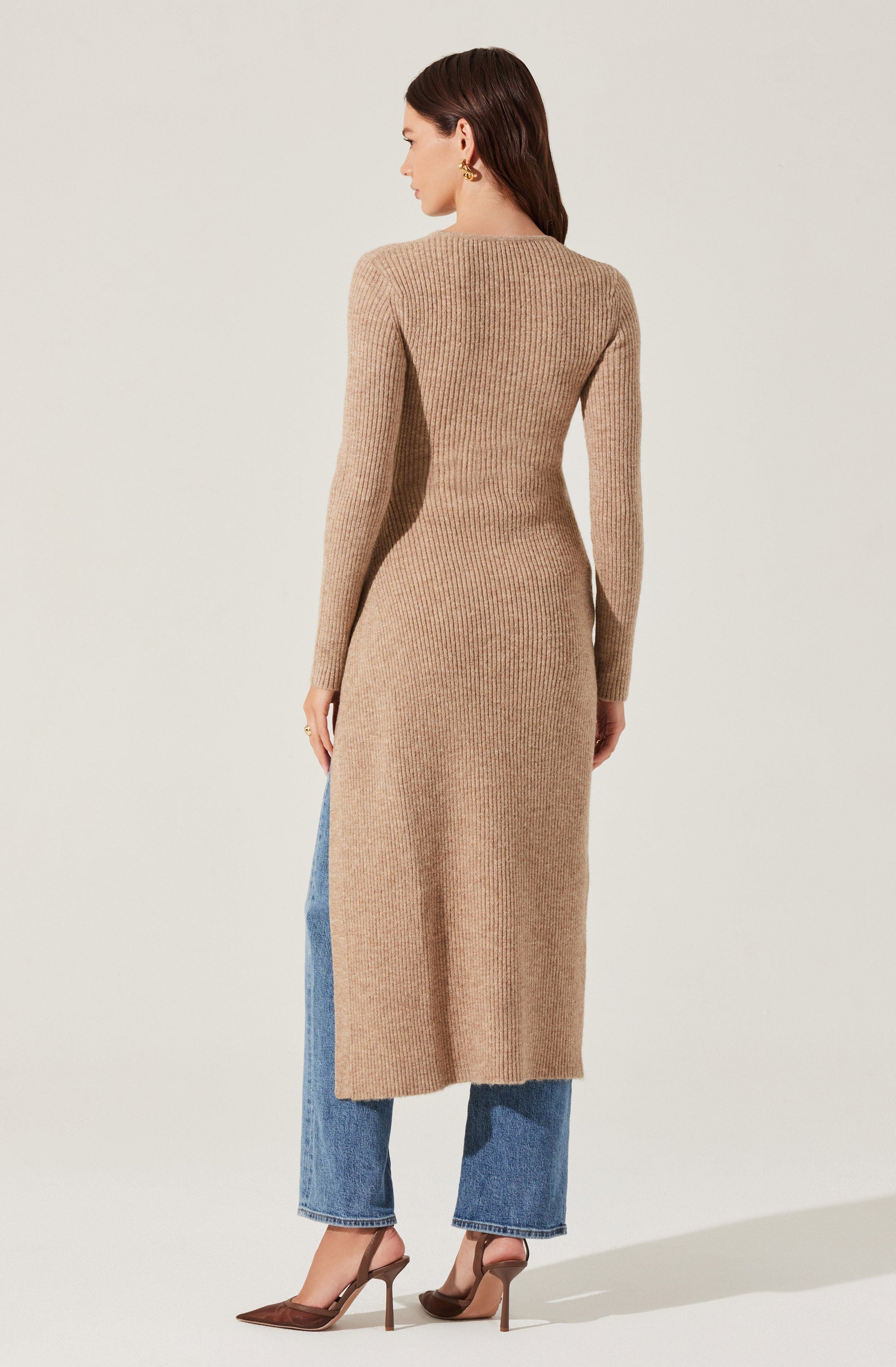 A woman is wearing a long, beige, ribbed knit sweater dress with long sleeves. She pairs it with medium-wash, wide-leg jeans that are cropped above the ankles. She is also wearing pointed-toe, high-heeled shoes in a brown shade.
