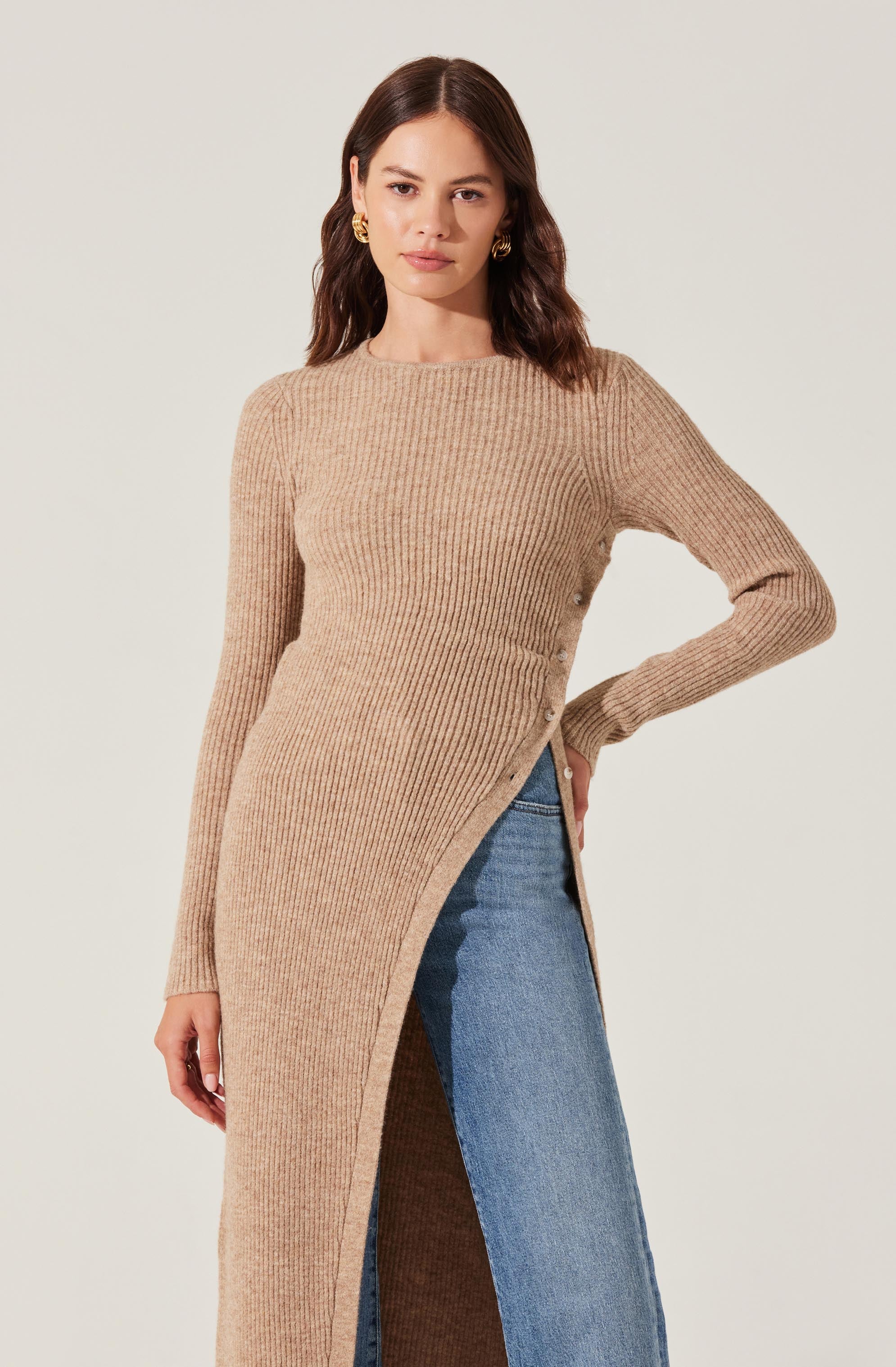 A woman is wearing a long, ribbed knit sweater in a beige color. The sweater features a high, round neckline, long sleeves, and side slits with buttons running along the edges. She has paired it with a blue denim skirt.