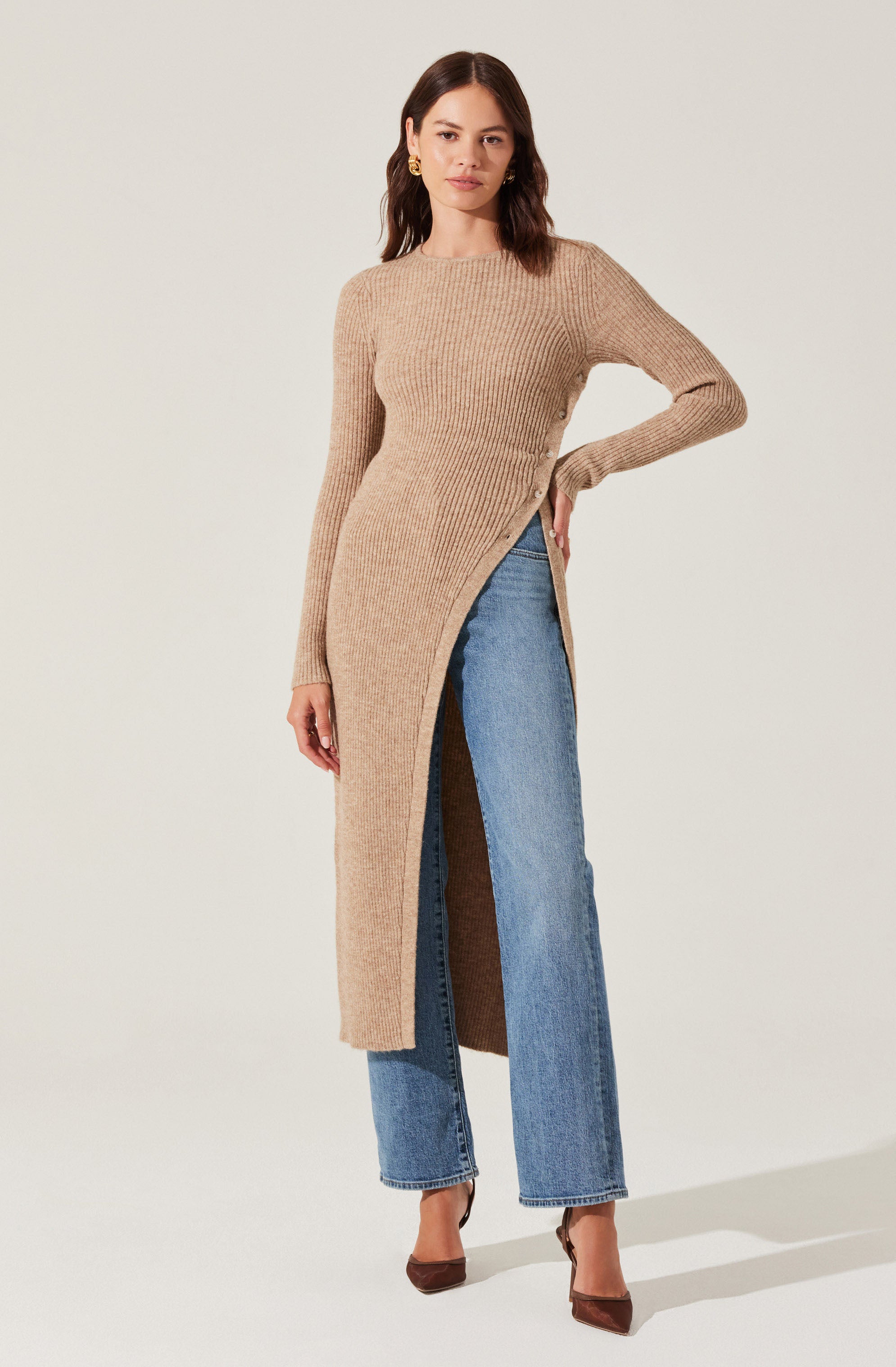 A woman is wearing a long, beige, ribbed knit sweater with a high neckline and long sleeves. The sweater has a front slit that reveals a pair of light blue, high-waisted, wide-leg jeans. The sweater's texture appears soft and cozy, with the slit adding a stylish detail. She is also wearing brown high heels.