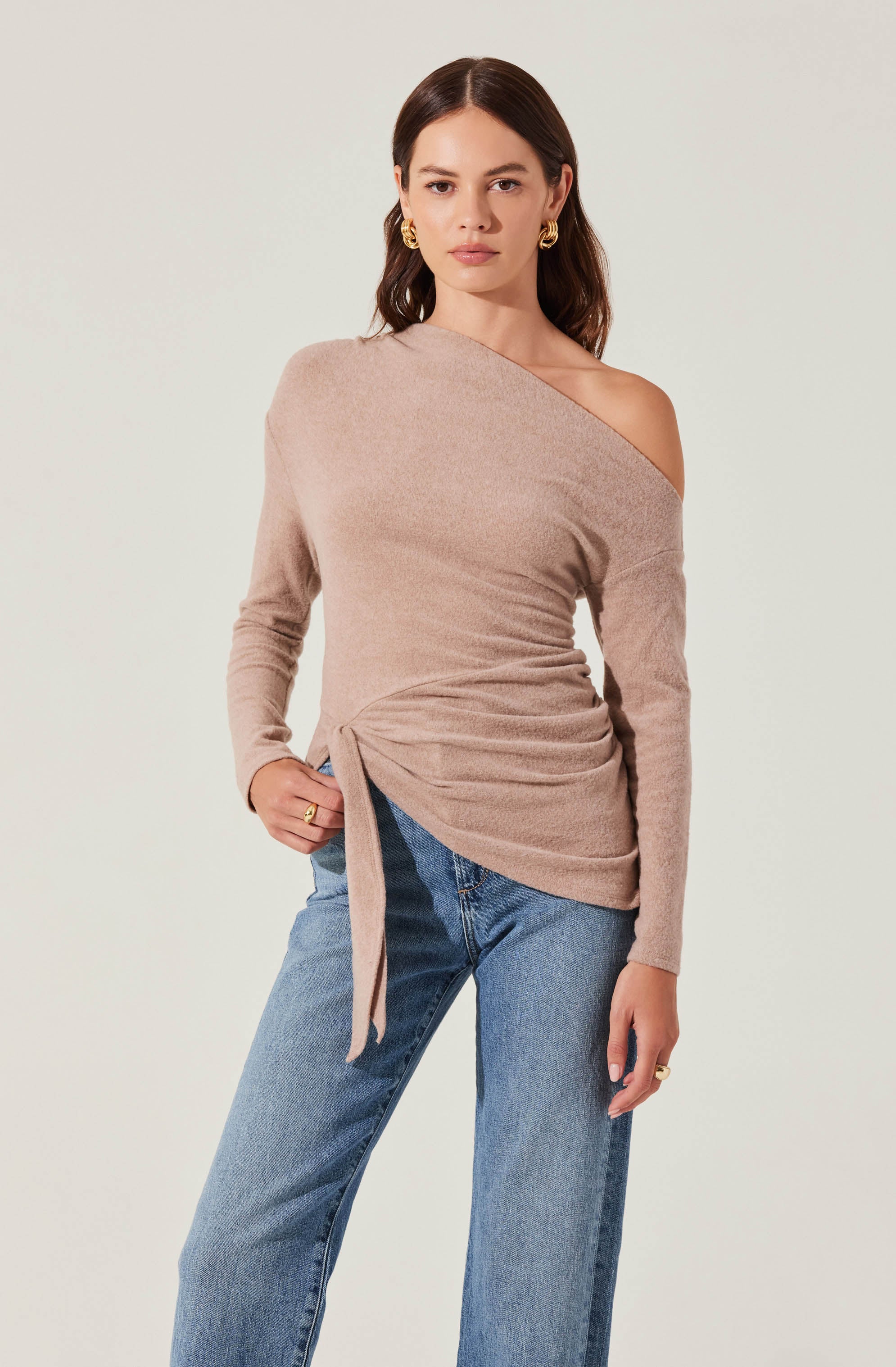 A woman is wearing a beige, off-shoulder sweater with a side tie detail, paired with light blue straight-leg jeans. She has dark brown hair, gold hoop earrings, and gold rings on her fingers.