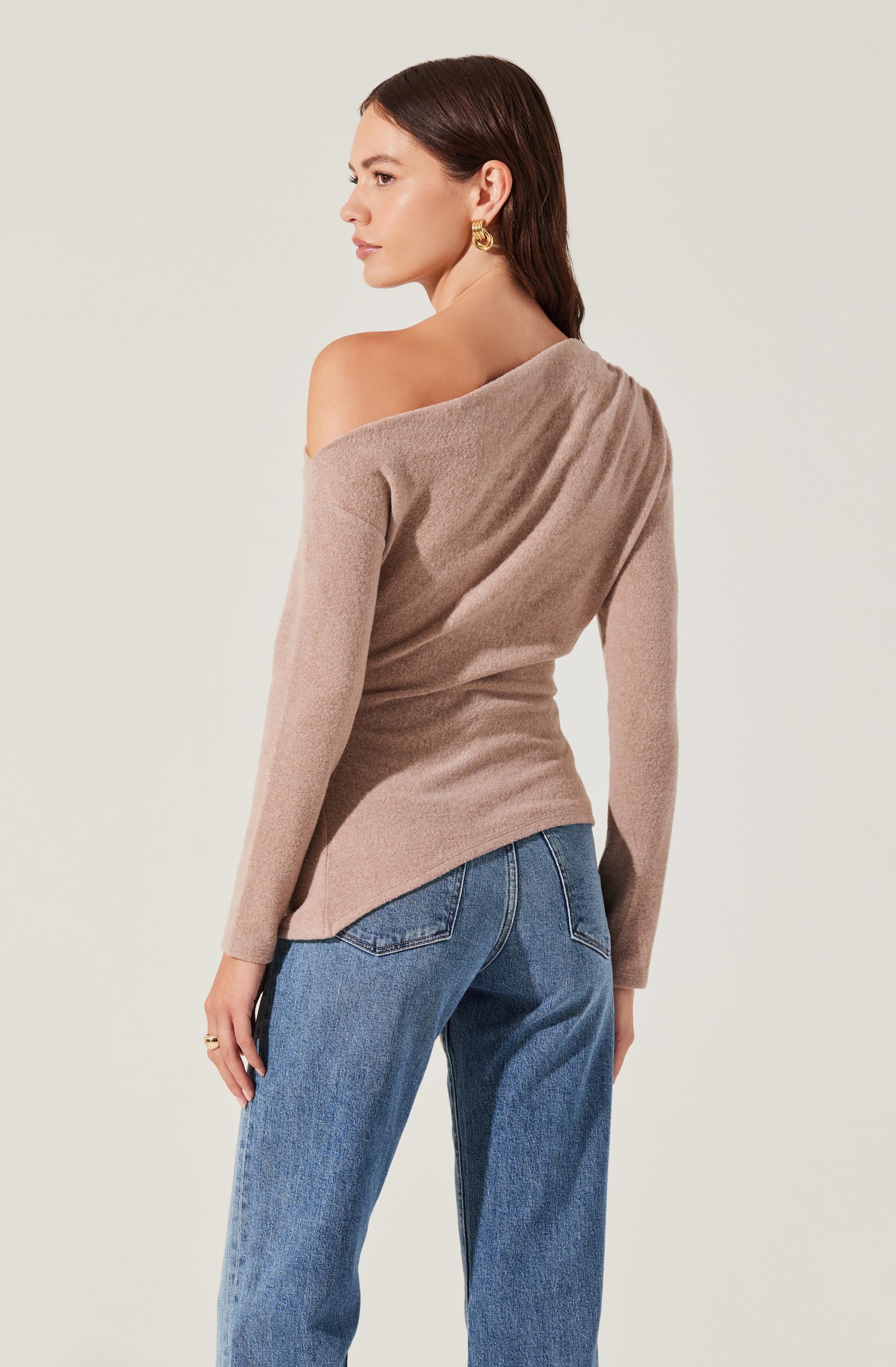 A woman is wearing a light beige, off-the-shoulder sweater with long sleeves, made from a soft, textured fabric. The sweater appears cozy and slightly loose-fitting. She pairs it with high-waisted, medium-wash blue jeans. She has shoulder-length dark brown hair and is accessorized with gold earrings.