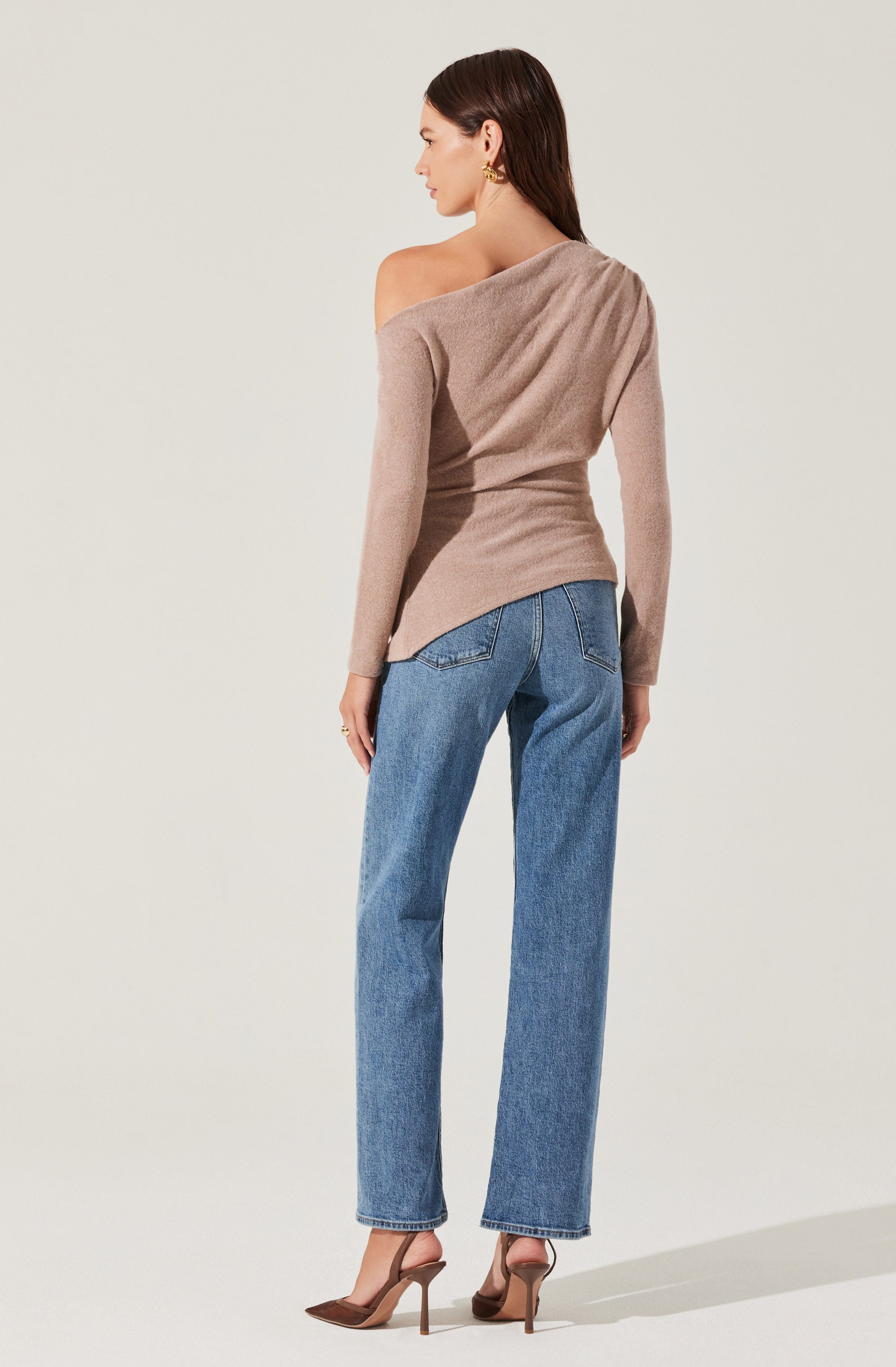 A woman is wearing a loose, off-the-shoulder beige sweater with long sleeves. She pairs it with high-waisted, straight-leg blue jeans and brown high-heeled shoes. She has gold earrings and her dark brown hair is styled straight.