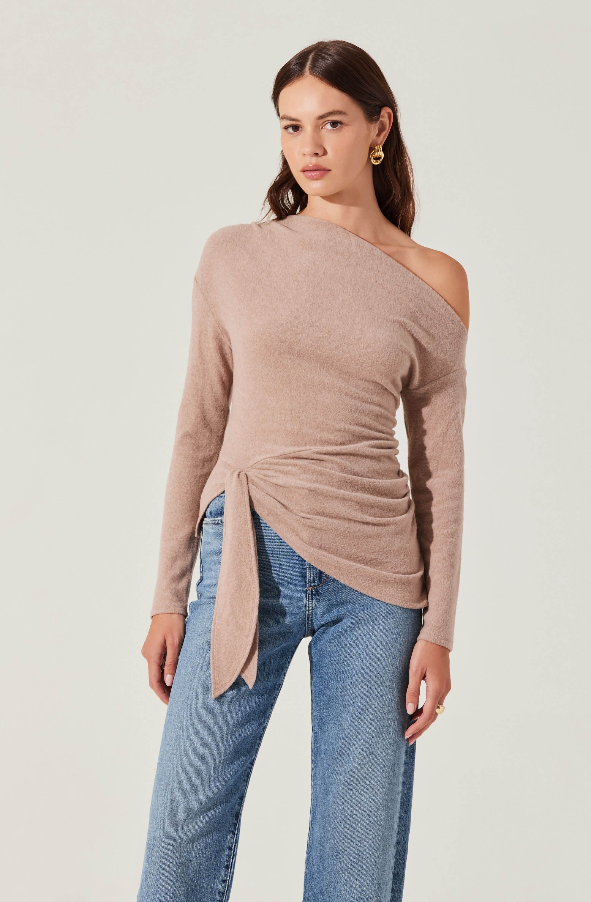 A woman is wearing a beige, long-sleeved, off-the-shoulder sweater with a draped, ruched detail on the side, tied in a bow. She pairs it with blue high-waisted jeans.