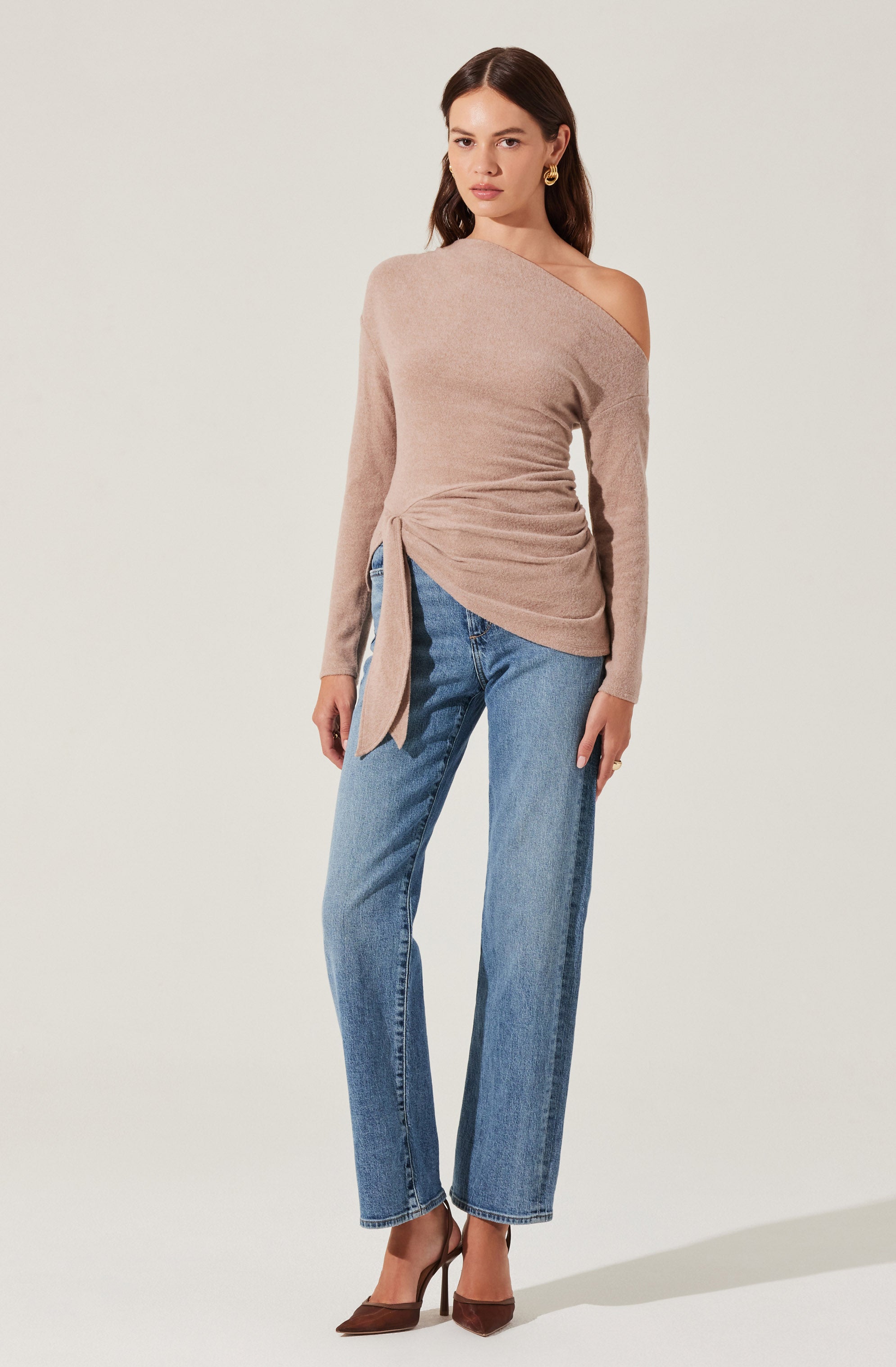 A woman is wearing a beige, one-shoulder sweater with a long sleeve on the left side and an asymmetrical neckline on the right side. The sweater features a ruched detail on the left side with a tie that hangs down. She pairs it with high-waisted, straight-leg blue jeans and brown high-heeled shoes.