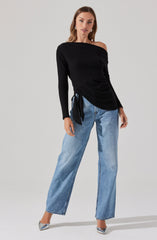 A woman is wearing a black off-the-shoulder long-sleeve top with a side tie detail. She is also wearing light-wash, wide-leg jeans. She has on silver pointed-toe high heels and is accessorized with earrings and rings.