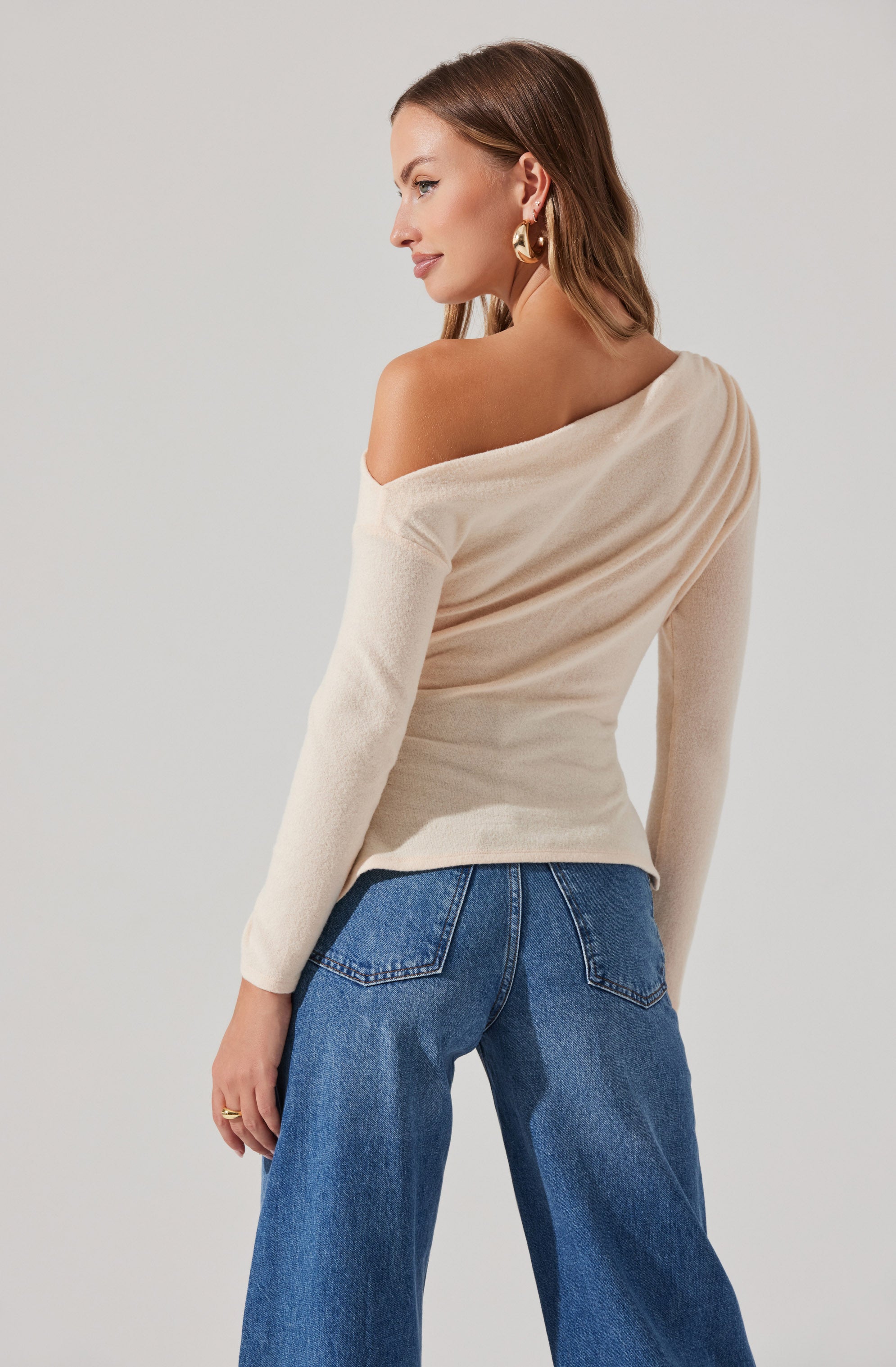 A woman is wearing a cream-colored, off-the-shoulder sweater with long sleeves. She is also wearing high-waisted, wide-leg blue jeans with visible back pockets.