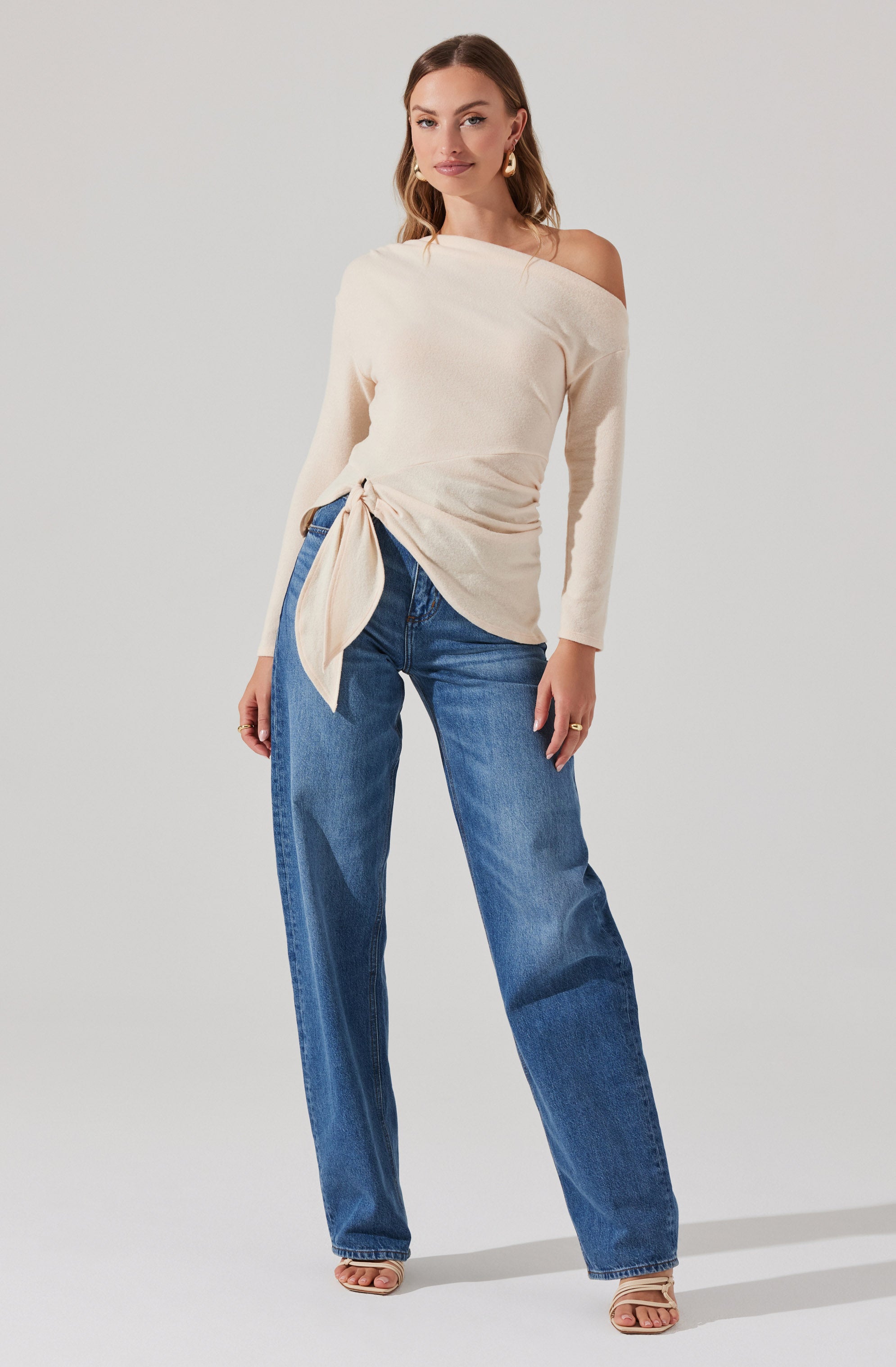 A woman is wearing a cream-colored, off-the-shoulder sweater with long sleeves. The sweater features a large tie detail at the waist on one side. She pairs it with high-waisted, relaxed-fit blue jeans and open-toe heels. She accessorizes with gold earrings and a ring.