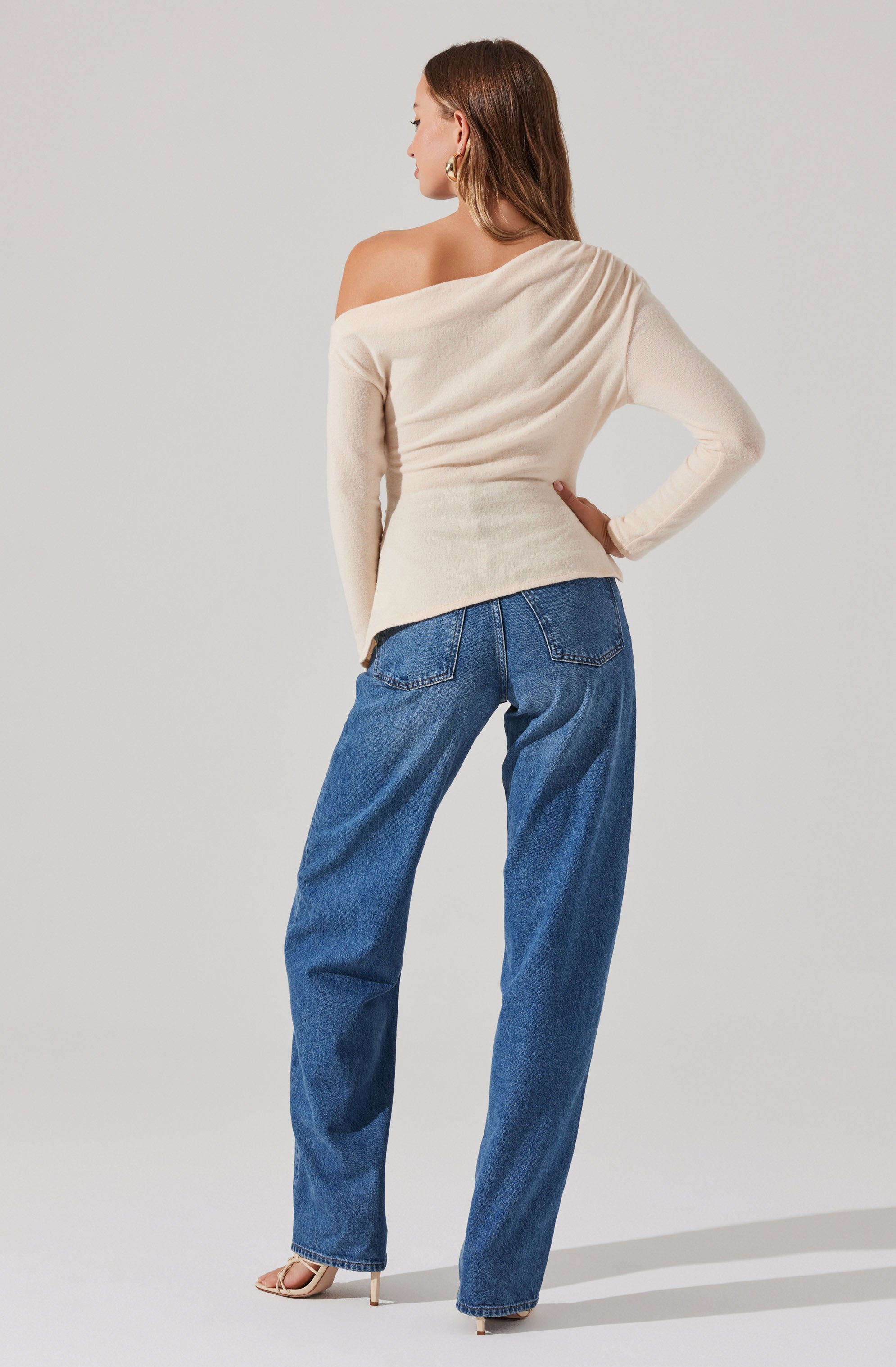A woman is wearing an off-the-shoulder, long-sleeve, beige sweater that drapes loosely on one side, creating an asymmetrical look. She pairs the sweater with high-waisted, loose-fitting blue jeans. Her shoes are pointed-toe heels with a thin ankle strap.