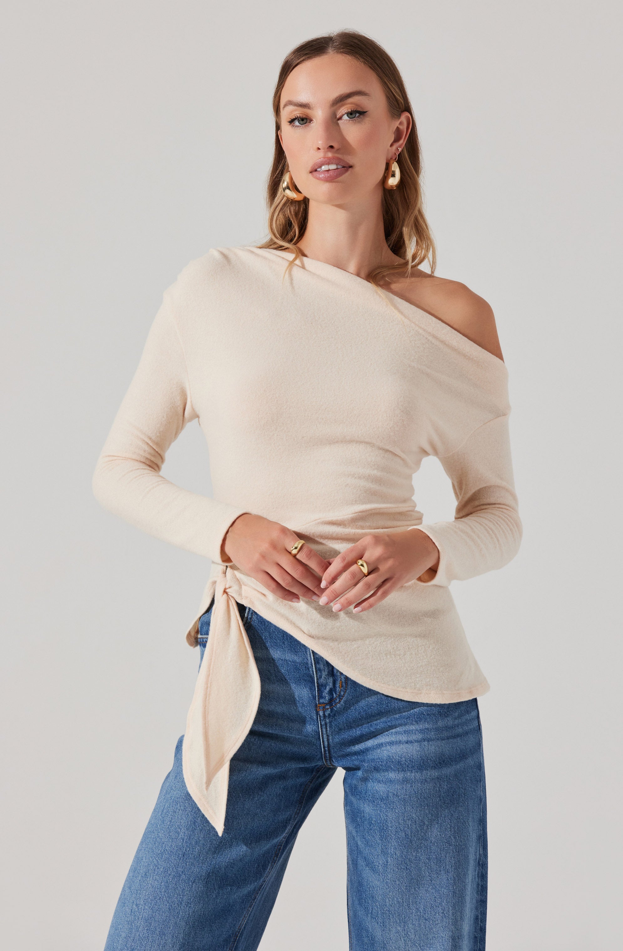 A woman is wearing a cream-colored, long-sleeve, off-the-shoulder top with a tie detail on one side. She has paired it with high-waisted blue jeans.