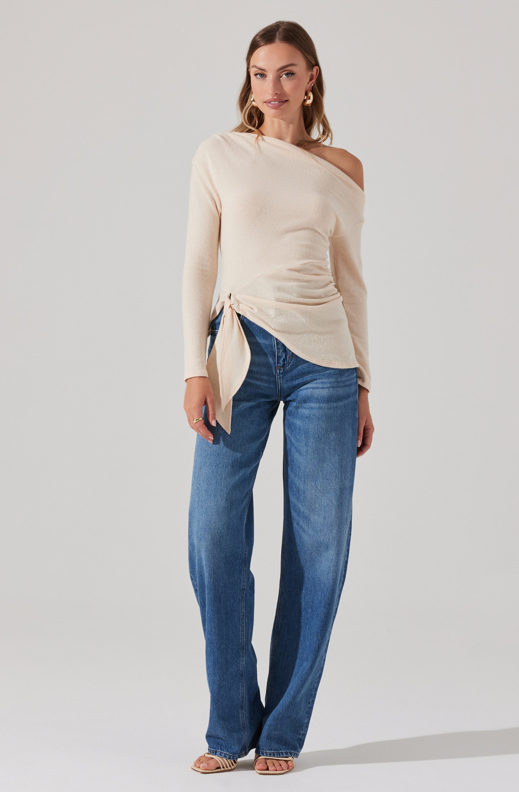 A woman is wearing a cream-colored, long-sleeved, off-the-shoulder top with a ruched detail and a tie on one side. She is also wearing high-waisted, straight-leg blue jeans.