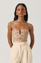A woman is wearing a strapless top with a leopard print pattern and a notch at the center of the neckline. She is also dressed in high-waisted, cream-colored trousers with pleats in the front. She has large, gold, hoop earrings and her hair is styled long and straight.