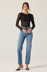 A woman is wearing a sheer black long-sleeve top with a round neckline. The top has a fitted bodice with vertical boning detail and is made of see-through fabric, revealing her skin underneath. She pairs this with high-waisted, straight-leg blue jeans and black open-toe high-heeled shoes.