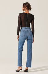 The image shows a woman wearing a fitted, long-sleeved black top made of sheer material, with a visible zipper running down the back. She is also wearing high-waisted, light blue jeans with a relaxed fit and classic five-pocket styling. She has on black high-heeled shoes and gold earrings. Her hair is styled in a low bun.