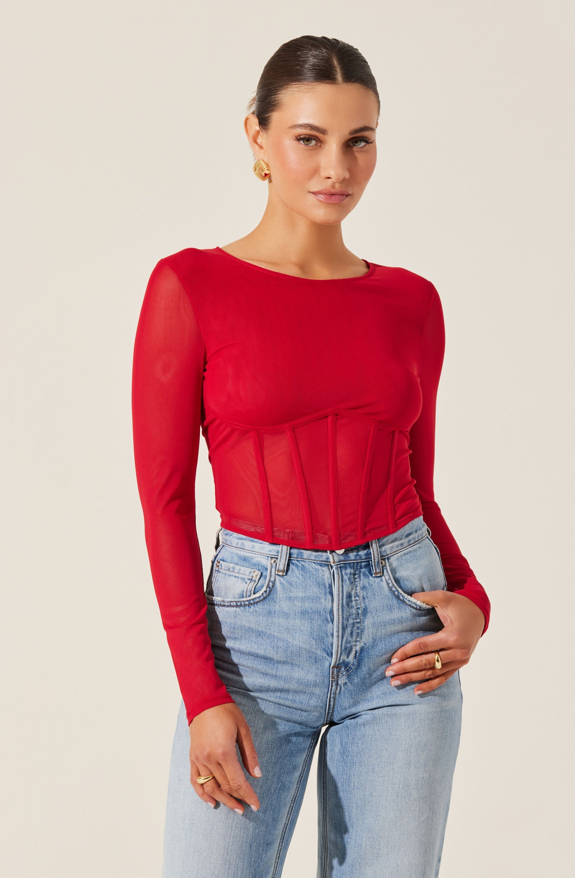 A woman is wearing a fitted, long-sleeved red top made of sheer, stretchy fabric. The top has a boat neckline and a structured corset-style design with vertical seams on the front. She pairs the top with high-waisted, light-wash jeans that feature dark side panels and a relaxed fit. She has gold earrings and rings on her fingers, and her dark hair is styled in a sleek, pulled-back look.