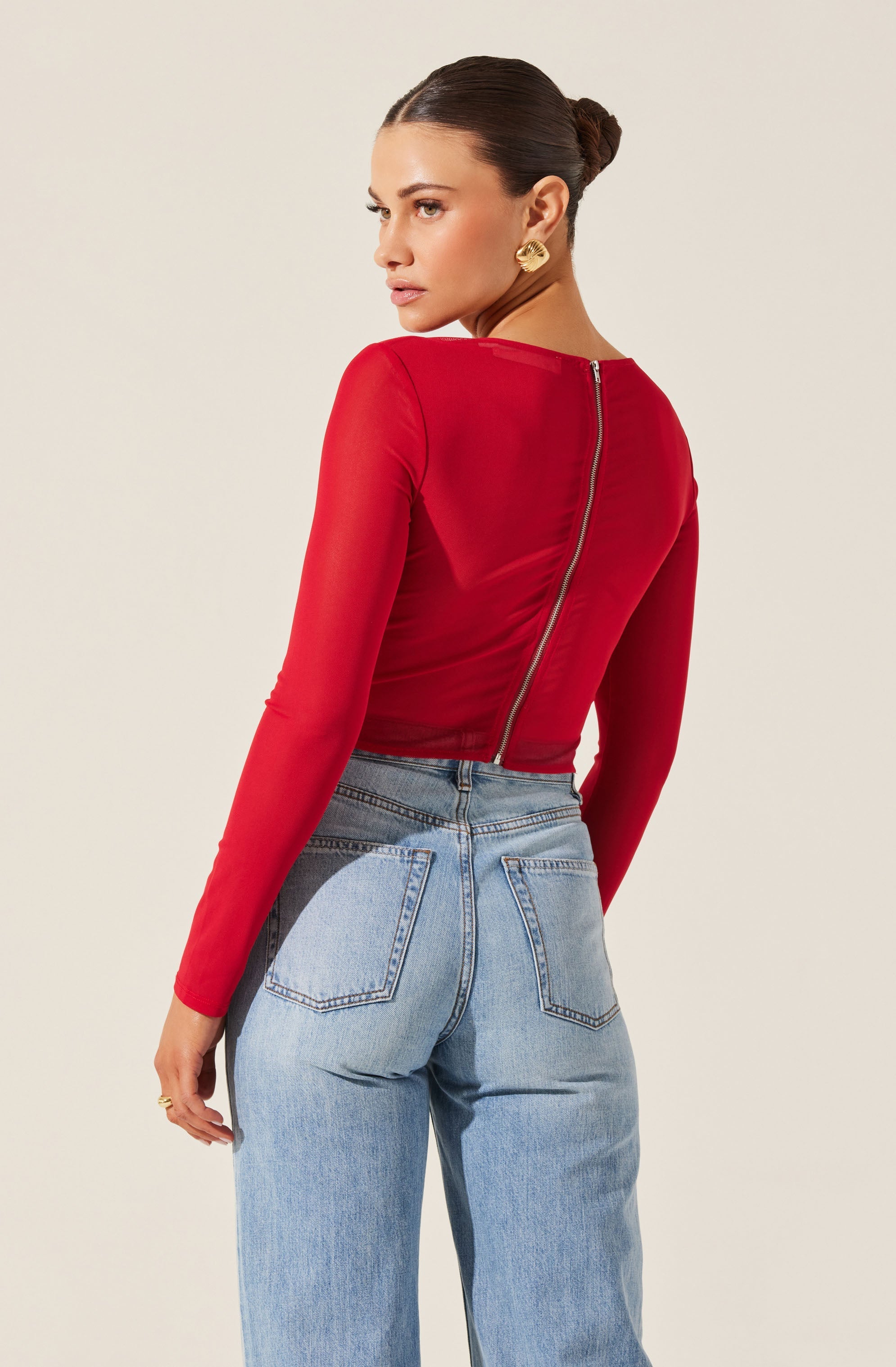 A woman is wearing a fitted, long-sleeved red top with a visible central zipper running from the neckline to the lower back. She pairs it with high-waisted, light-wash denim jeans that have prominent back pockets and contrast stitching. The top has a smooth, slightly shiny texture, and the jeans have a relaxed fit.