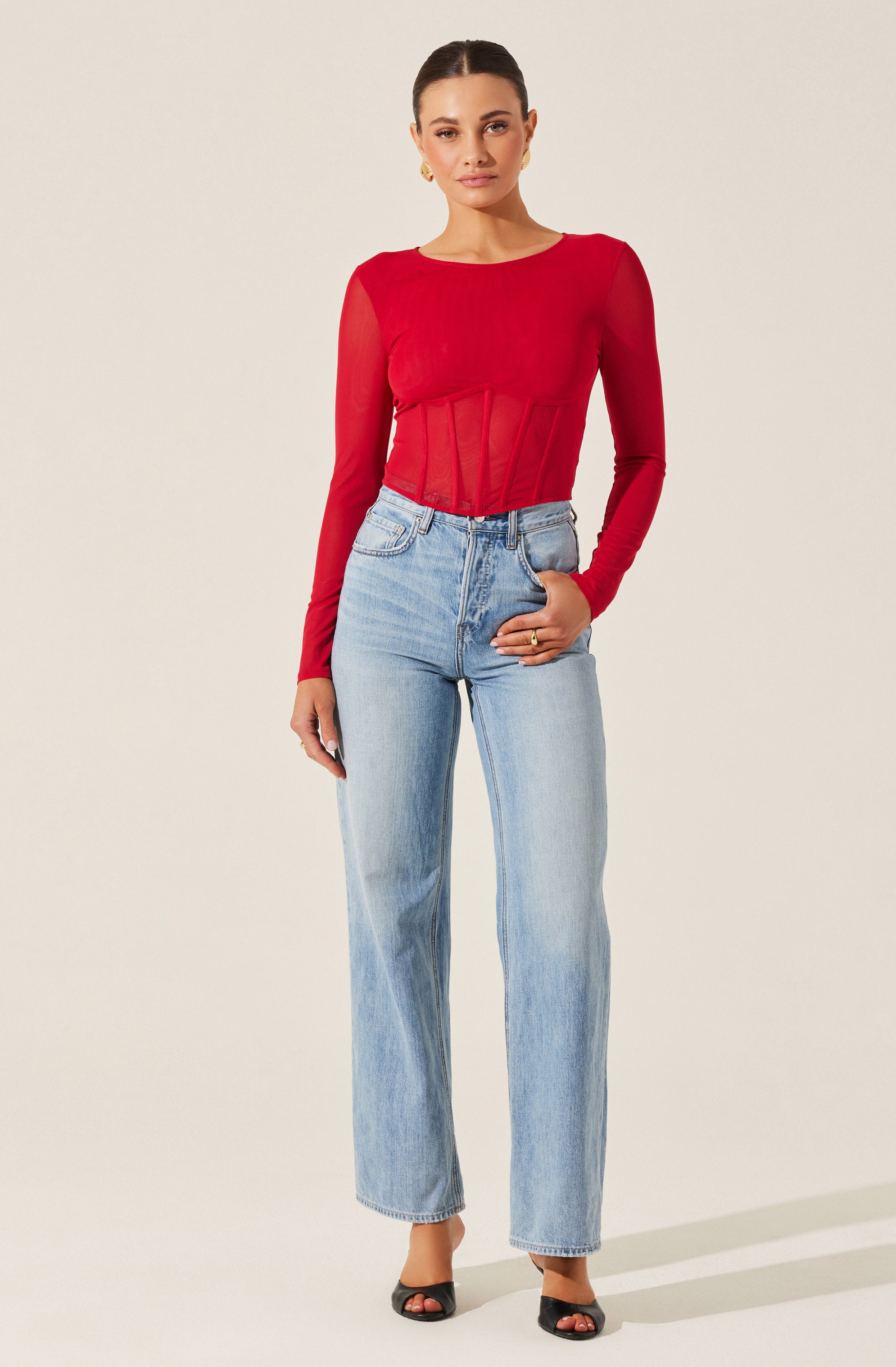 A woman is wearing a fitted, long-sleeved red top with a round neckline. The top features sheer, mesh-like details in the front with vertical paneling. She is also wearing high-waisted, light-wash straight-leg jeans. She has on black open-toe heels and is accessorized with gold earrings and a gold ring.