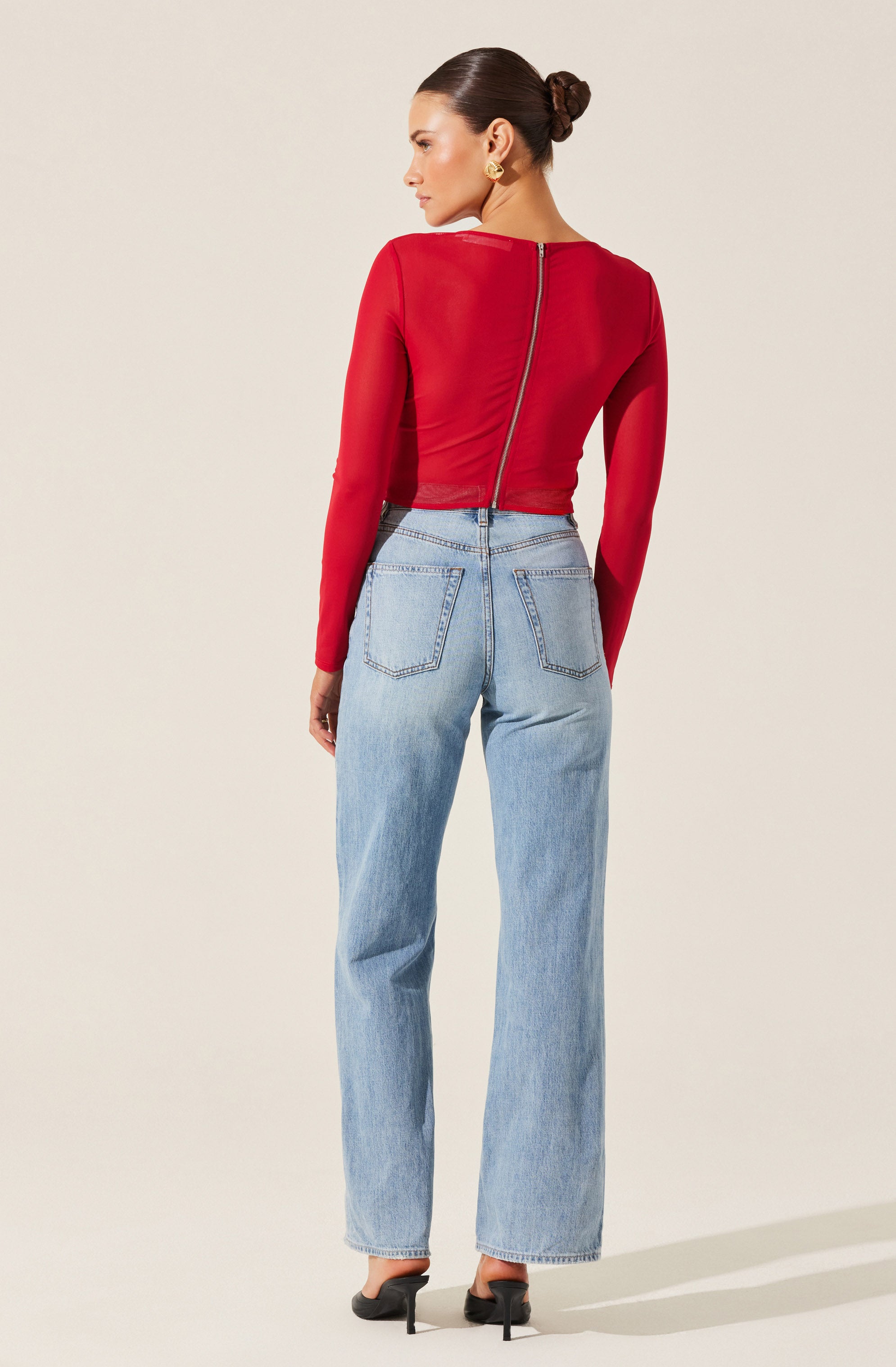 A woman is wearing a fitted, long-sleeve red top with a diagonal zipper running from the back neckline to the side waist. She pairs it with high-waisted, light-wash denim jeans that have prominent back pockets and a wide-leg cut. She is also wearing black high-heeled shoes.