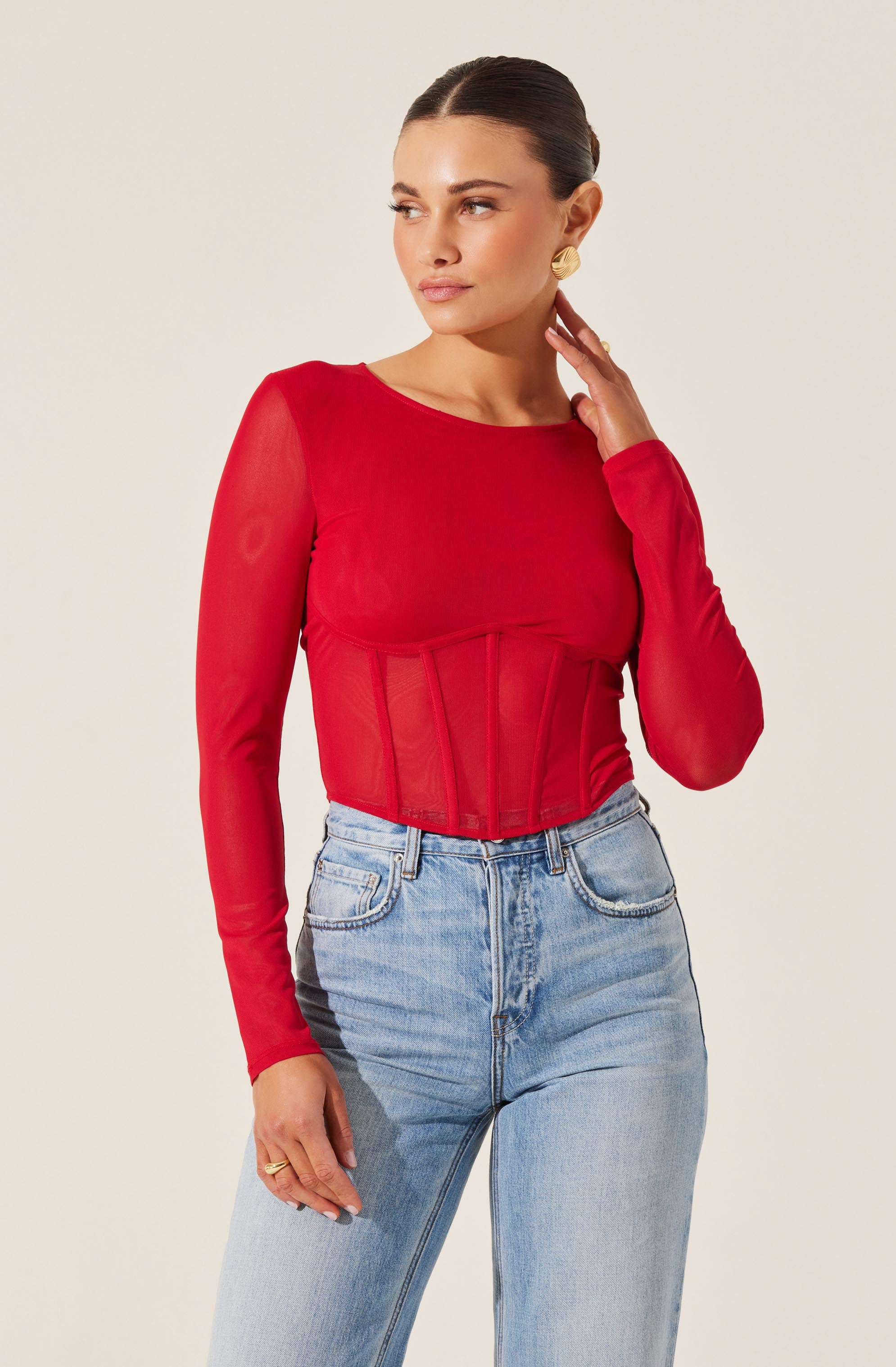 A woman is wearing a fitted, long-sleeved, red top with sheer sleeves. She is also wearing high-waisted, light-wash jeans. The outfit is accessorized with large, circular gold earrings and a ring on her left hand.