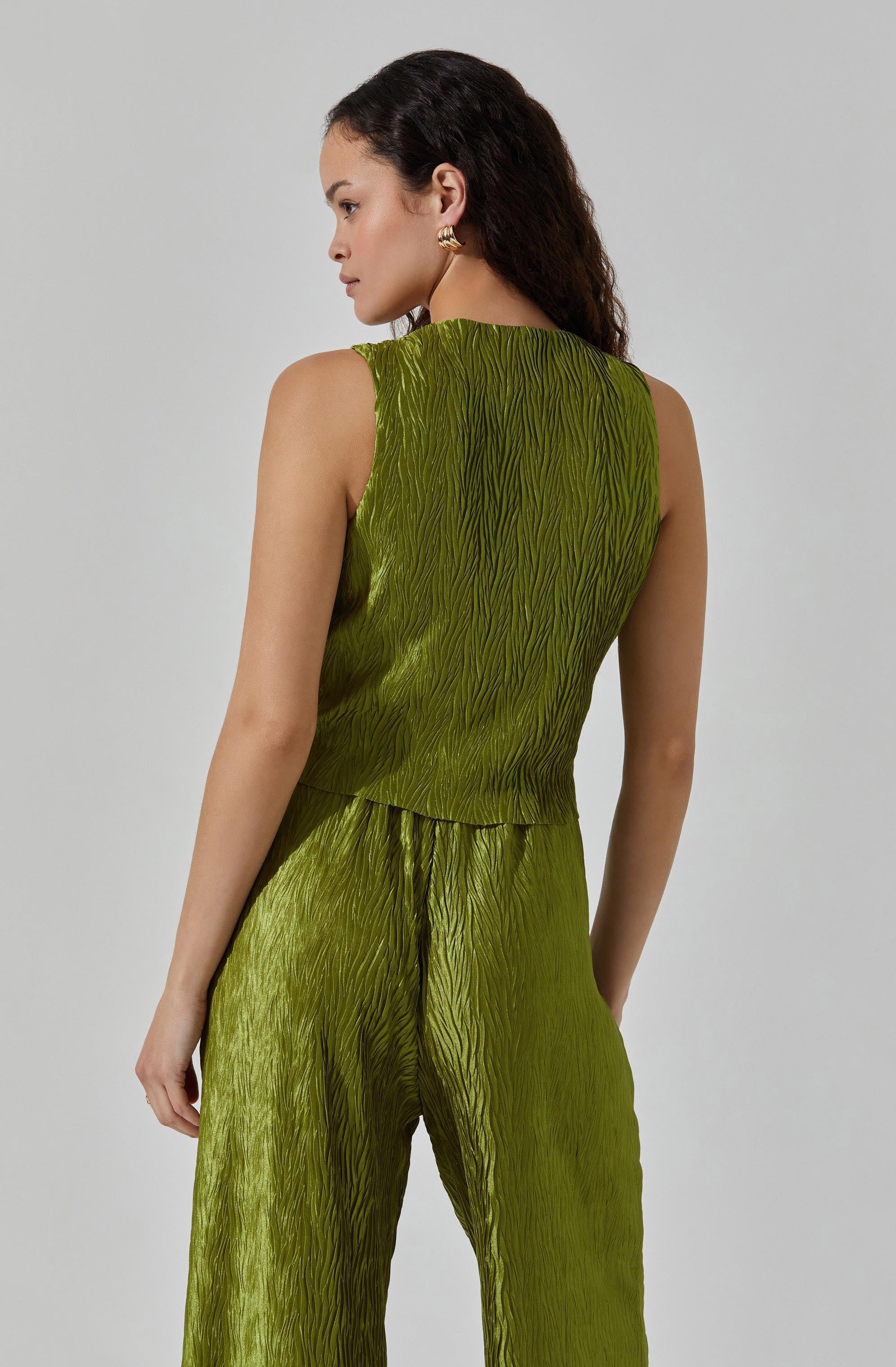 A woman is wearing a sleeveless, matching olive green set consisting of a top and wide-leg pants. The fabric has a textured, crinkled appearance with a subtle sheen. The top is fitted at the waist, while the pants have a relaxed fit with a slightly high waist. She has dark, wavy hair and is wearing gold earrings.