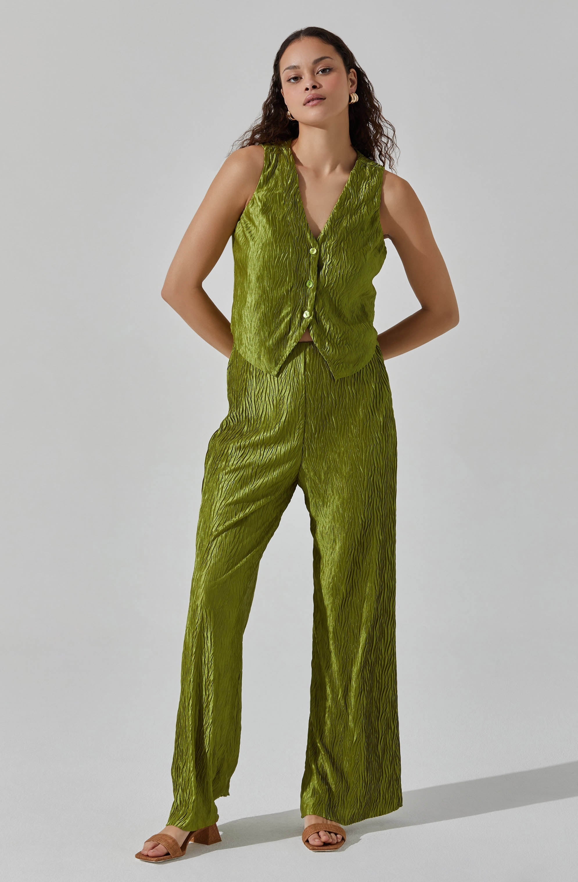 A woman is wearing a sleeveless, V-neck, button-up top and matching wide-leg pants made from a shiny, textured fabric in a bright lime green color. The fabric has a crinkled, vertical pattern. She completes the look with open-toe sandals.