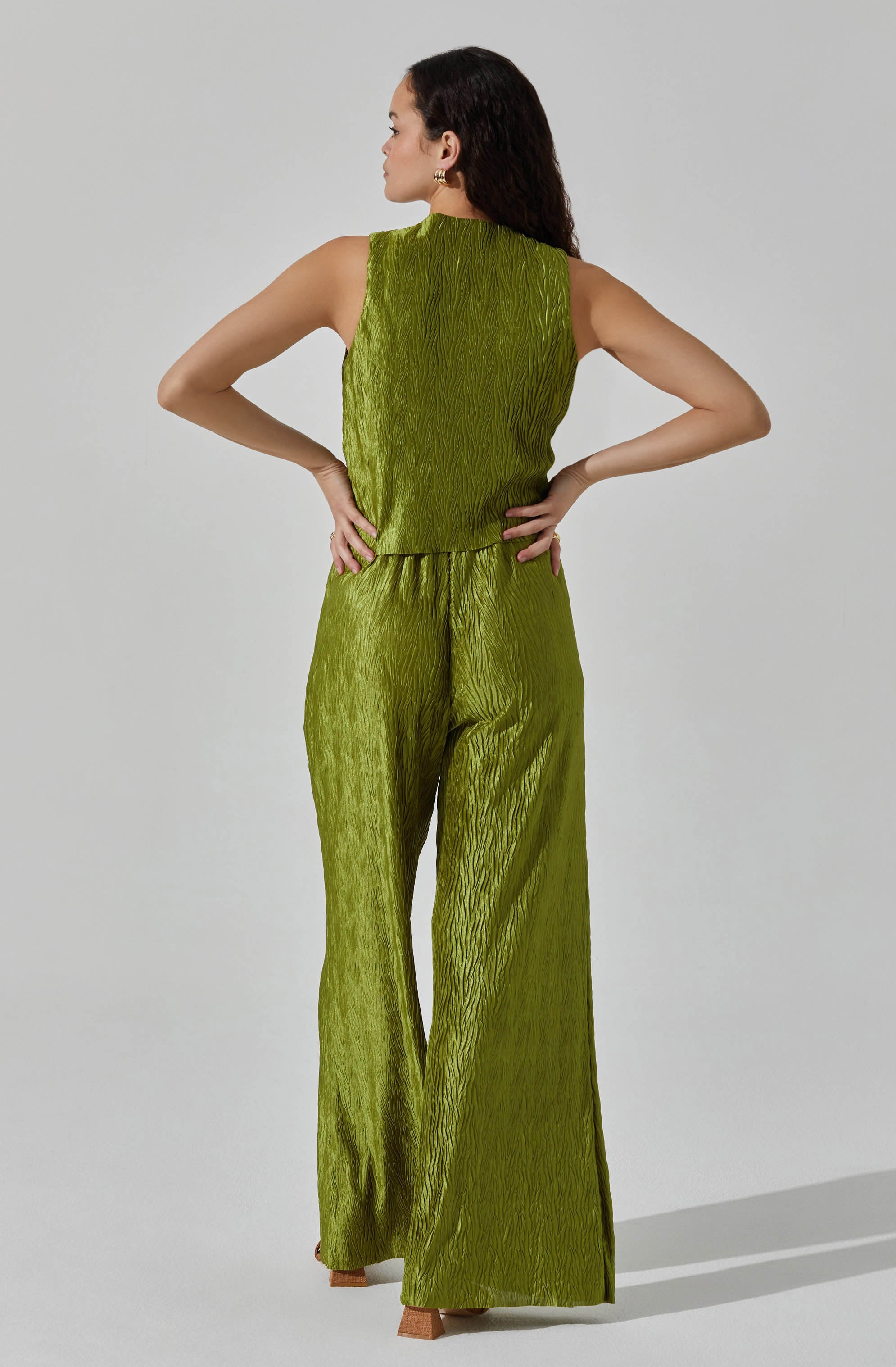 A woman is wearing a sleeveless, olive-green pantsuit with a textured, pleated fabric. The top has a high neckline and a loose fit, while the wide-leg pants feature the same textured, pleated design throughout.