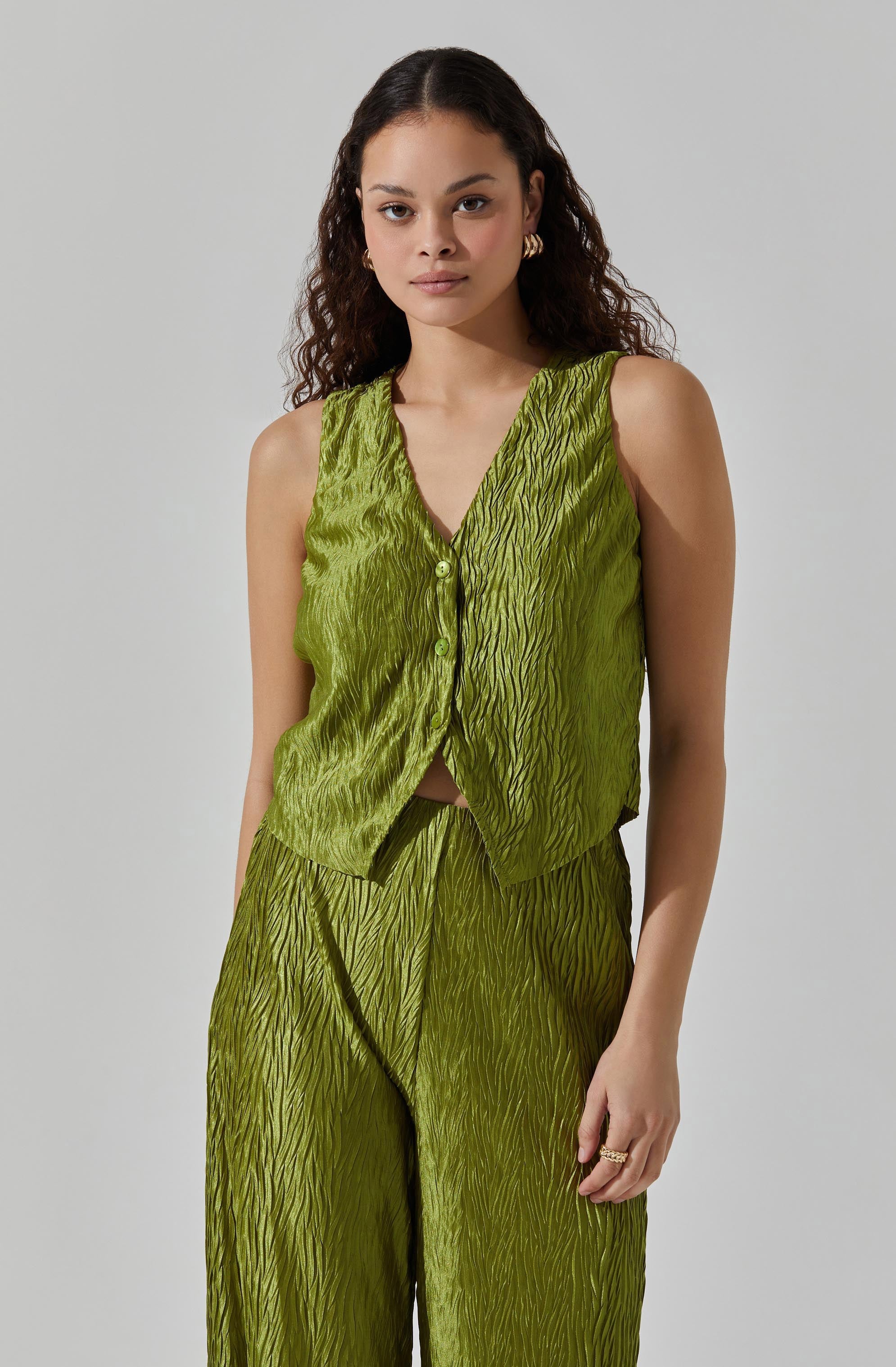 A woman is wearing a sleeveless, lime green outfit made of textured, shiny fabric with a wave-like pattern. The top is a vest style with a V-neck and buttons down the front, paired with matching wide-leg pants featuring the same textured design.
