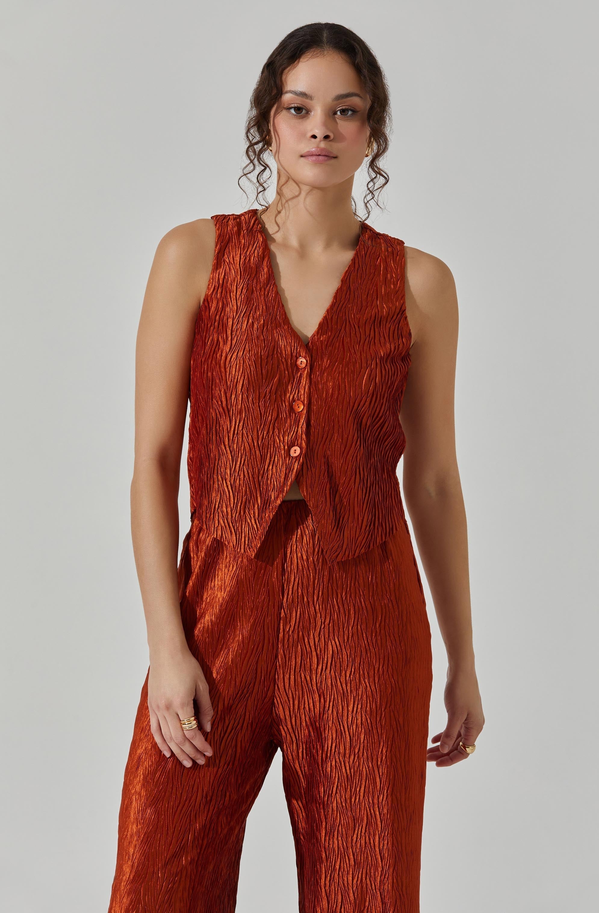 A woman is wearing a sleeveless, button-up vest and matching wide-leg pants. The fabric has a textured, crinkled appearance with a shiny finish, in a rich, burnt orange color.