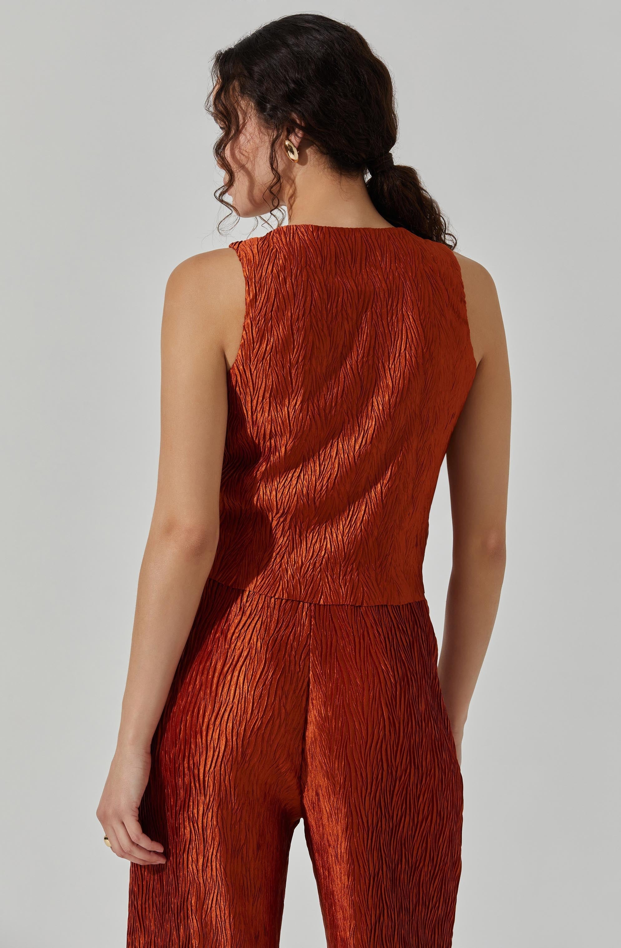 The image shows a person wearing a sleeveless, rust-colored outfit made from a textured, crinkled fabric. The top is fitted and has a high, round neckline, while the matching bottoms are high-waisted with a relaxed fit. The fabric appears shiny, giving the outfit a luxurious appearance. The person's dark, curly hair is styled in a loose ponytail, and they are accessorized with small gold hoop earrings.